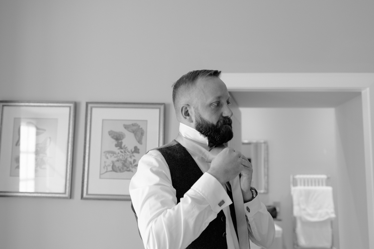black and white photo of groom in white shirt and waistcoat tying his tie in room scottish humanist wedding
