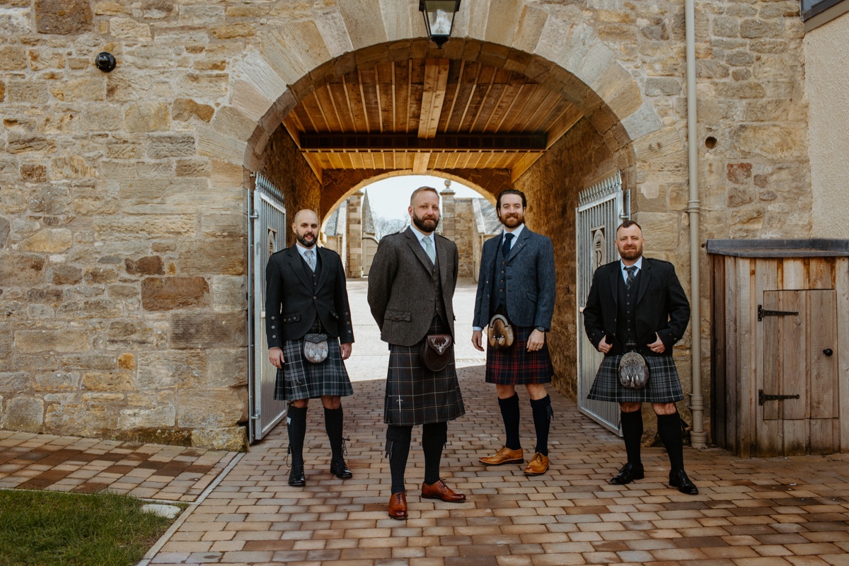 groom and best men wearing kilt outfits standing in front of stone arch at steadings rosebery steadings wedding