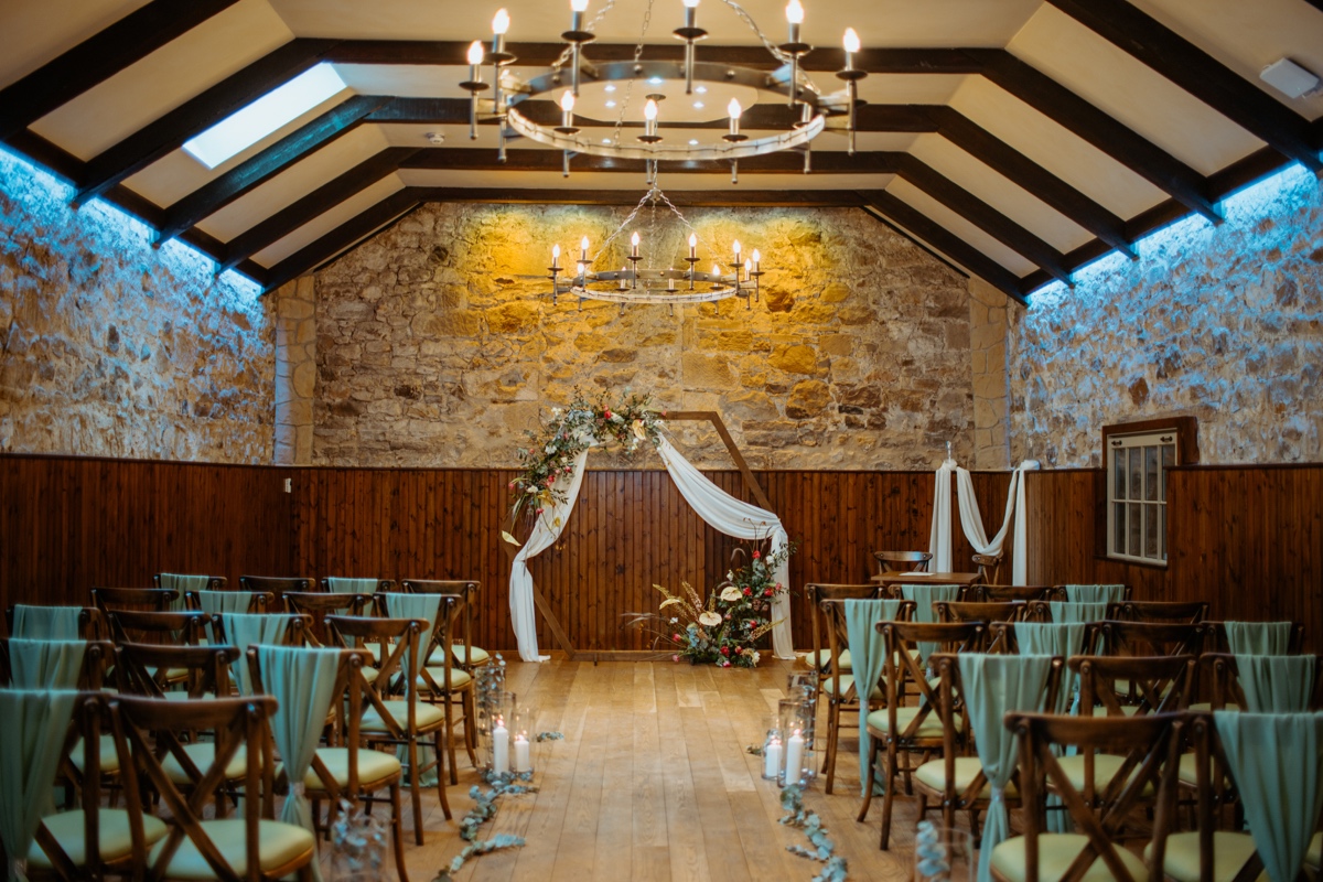 view of ceremony location in stone walled room in rosebery steadings wooden seats at each side chandeliers hanging from ceiling scottish humanist wedding
