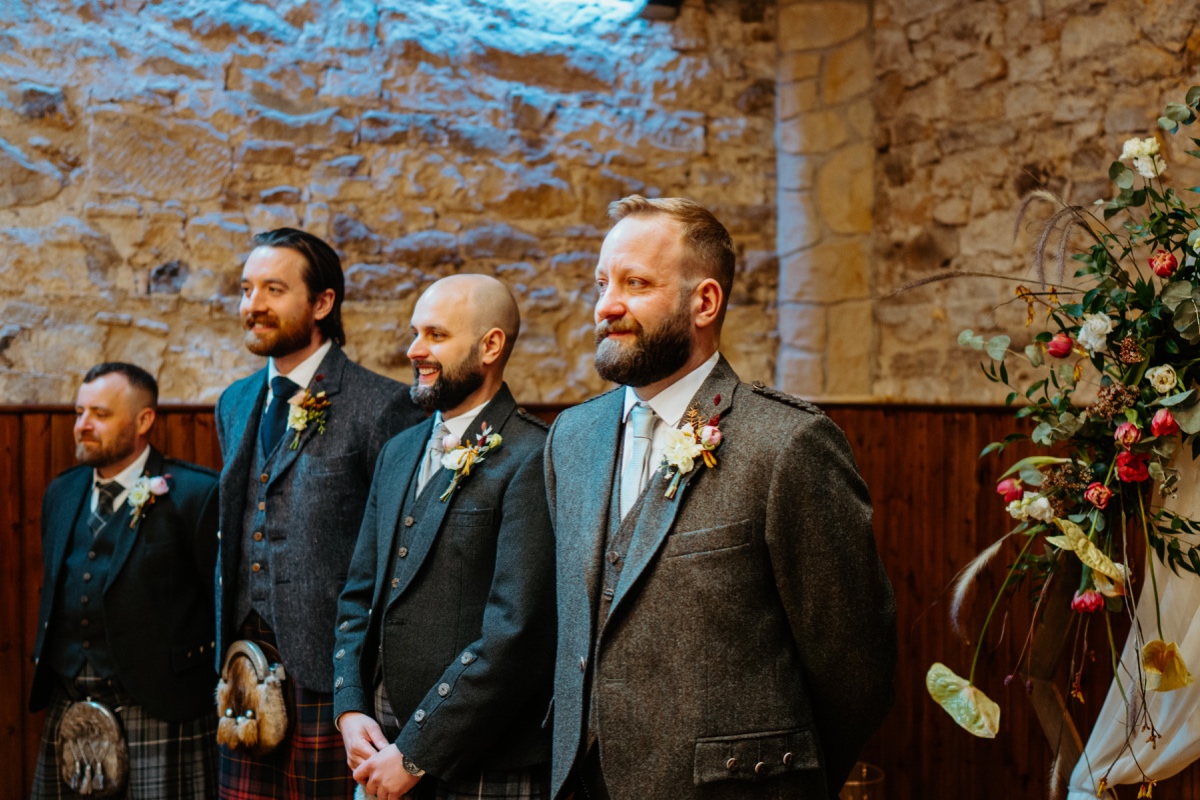 groom and best men smiling as they wait for the bride to walk down the aisle they are all wearing tweed jackets with stone walls behind them  scottish humanist wedding