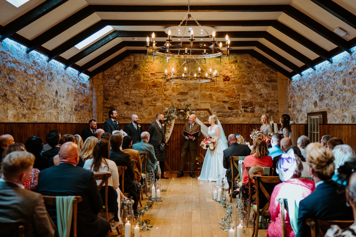 view of the ceremony from the back in stone walled room of steadings bride and groom facing each other as the guests look on scottish wedding photography rosebery steadings wedding