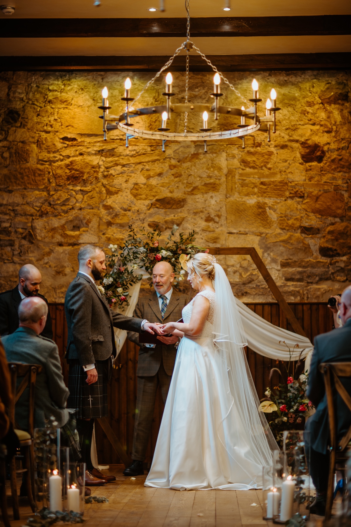 bride wearing white dress placing the ring on grooms finger with floral arch and stone wall in background scottish humanist wedding