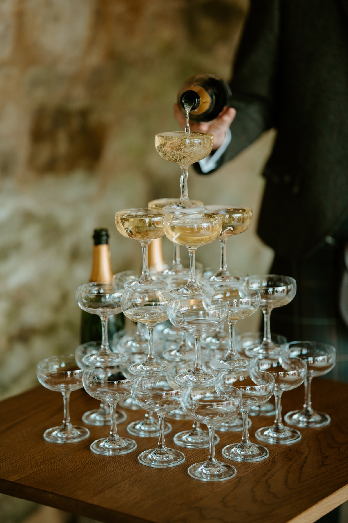 groom pouring champagne on to tower of champagne glasses on wooden table Humanist scotland rosebery steadings wedding