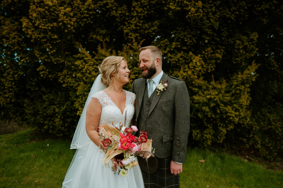 bride wearing white dress and groom wearing kilt outfit looking lovingly into each others eyes on grounds of rosebery steadings with green trees in background scottish humanist wedding