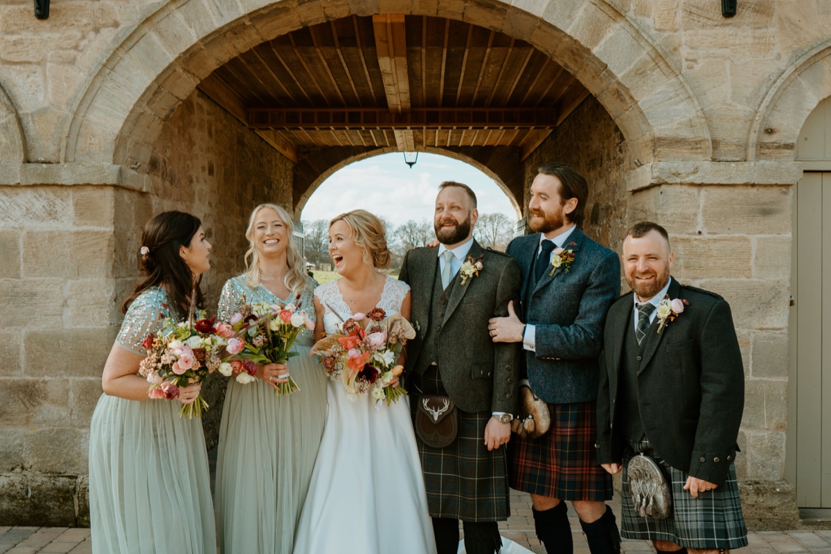 bride and groom posing and laughing with wedding party in front of stone arch at rosebery steadings scottish humanist wedding