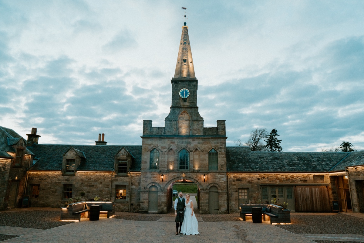 bride and groom posing in courtyard of of rosebery steadings with clock tower behind them scottish humanist wedding