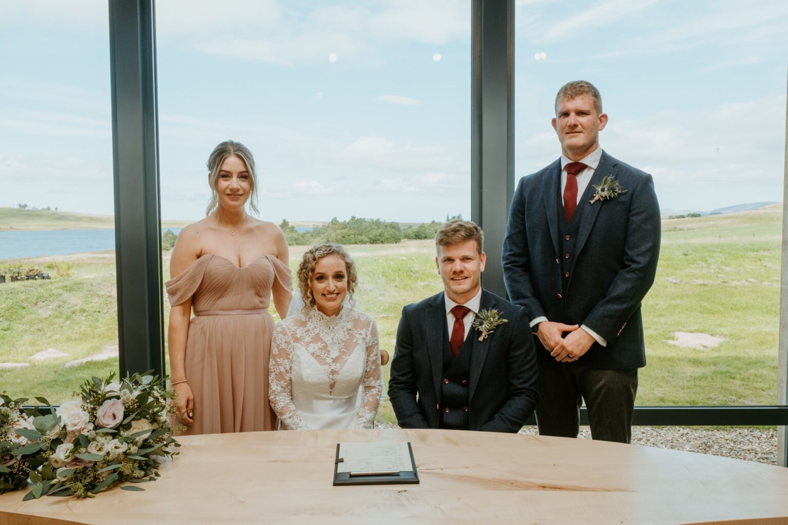 bride and groom sitting at table with best man and bridemaid standing behind them big glass window behind them showing view of fields and loch cairns farm estate wedding