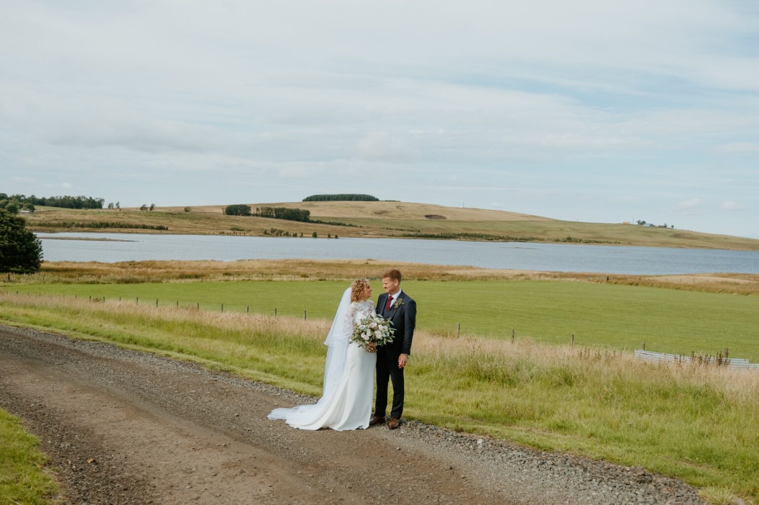 bride and groom looking into the eyes of each other as they stand on country road with loch and green hills in background cairns farm estate wedding