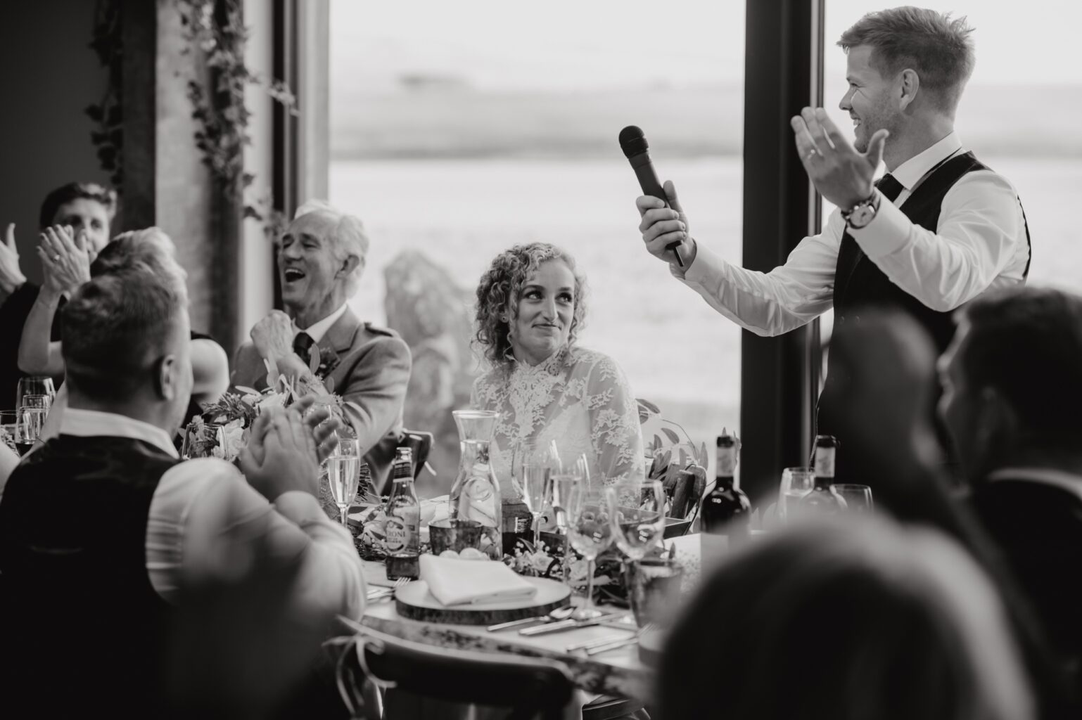 black and white photo of groom making speech with guests clapping and laughing cairns farm estate wedding