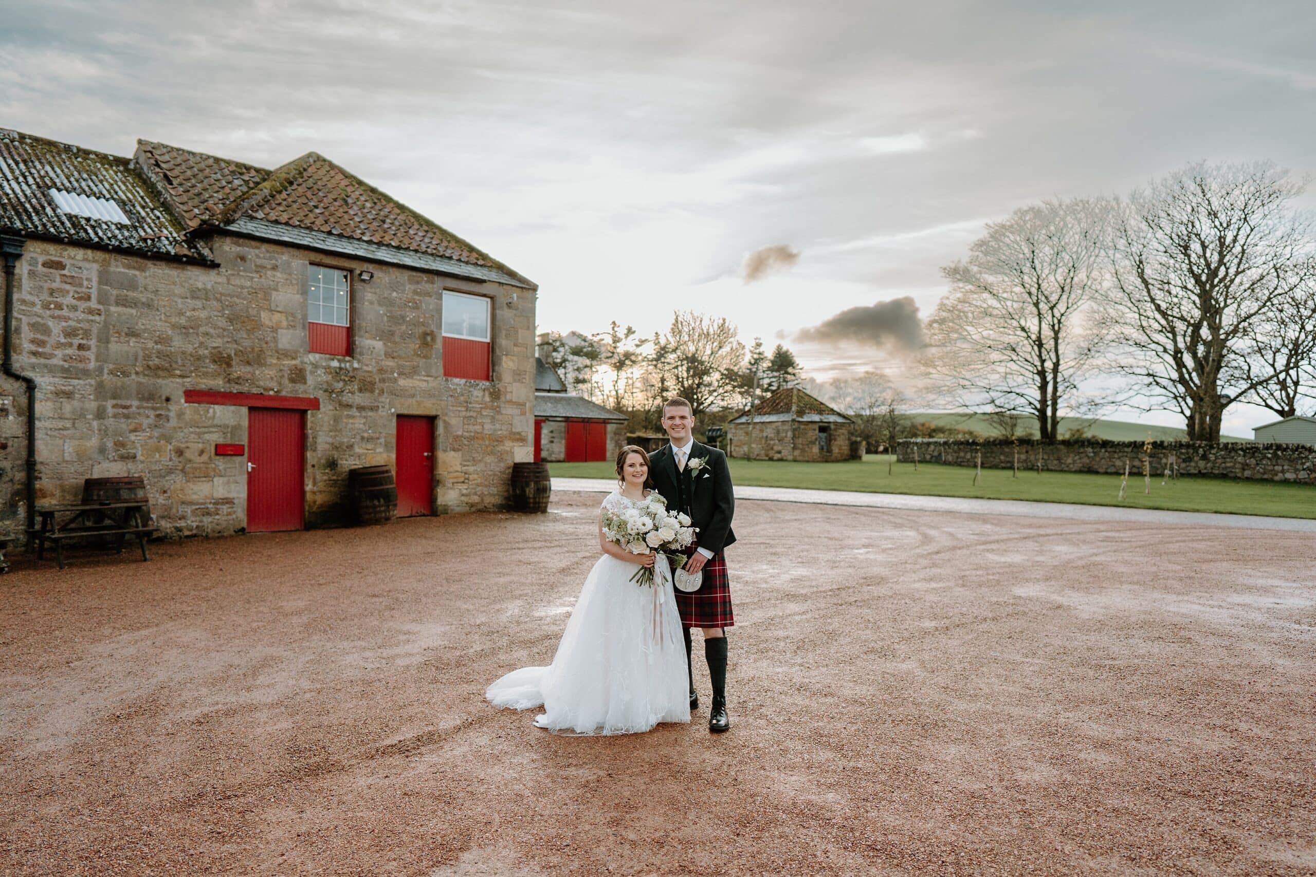 kinkell byre wedding photos of bride and groom outside venue with barn wedding venue st andrews in the background