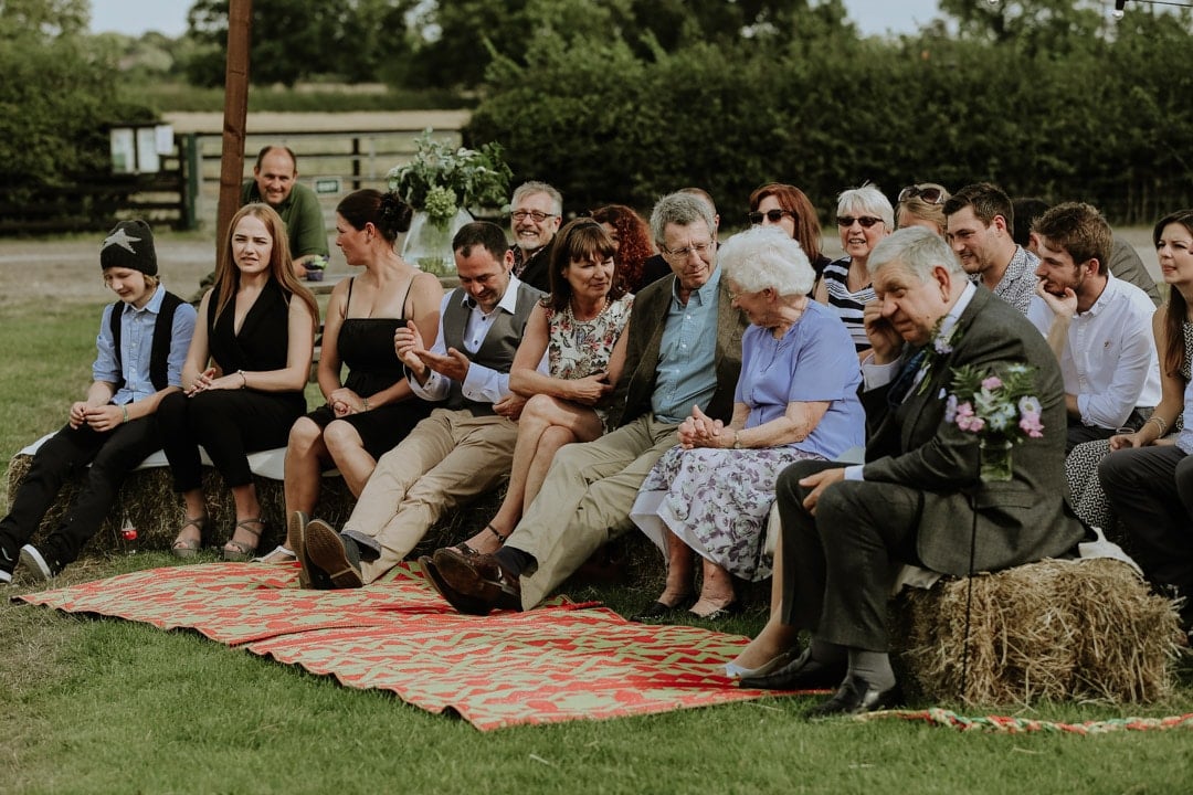 guests-sit-upon-haybales-awaiting-bride-rugs-outdoors-wedding