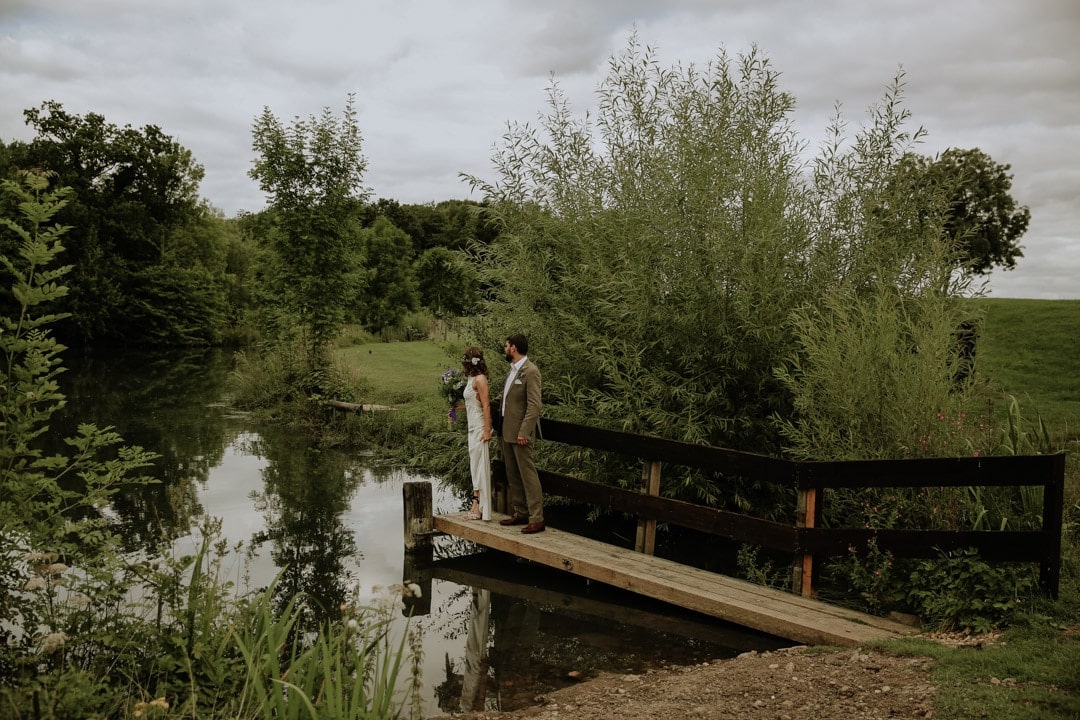bride-stands-with-groom-holding-hands-countryside-lake-diy-wedding