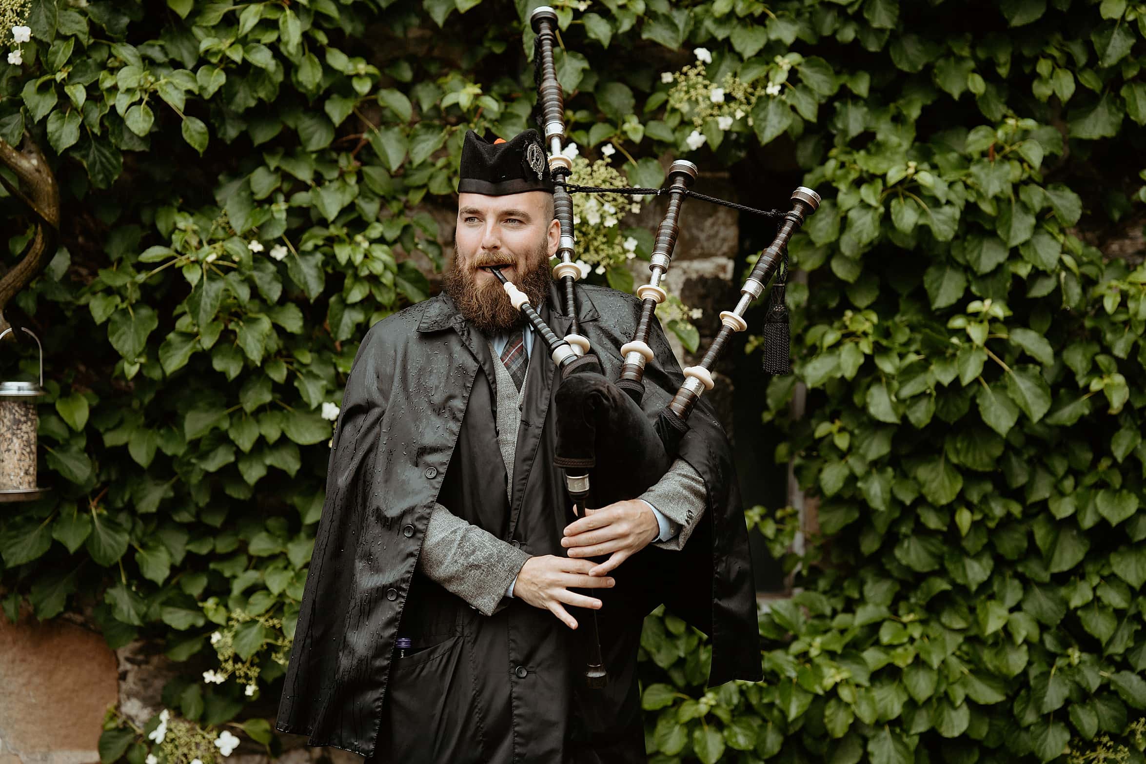 bagpiper wearing black hat and black raincoat piping in front of ivy wall monachyle mhor wedding