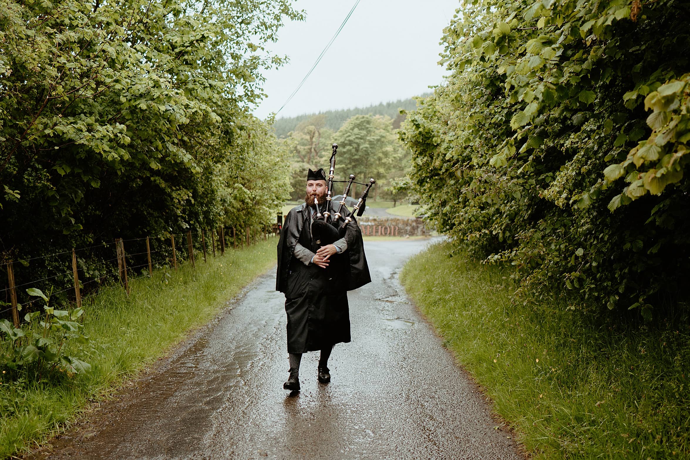 bagpiper walking towards camera on country road in rain with trees at either side of road monachyle mhor wedding