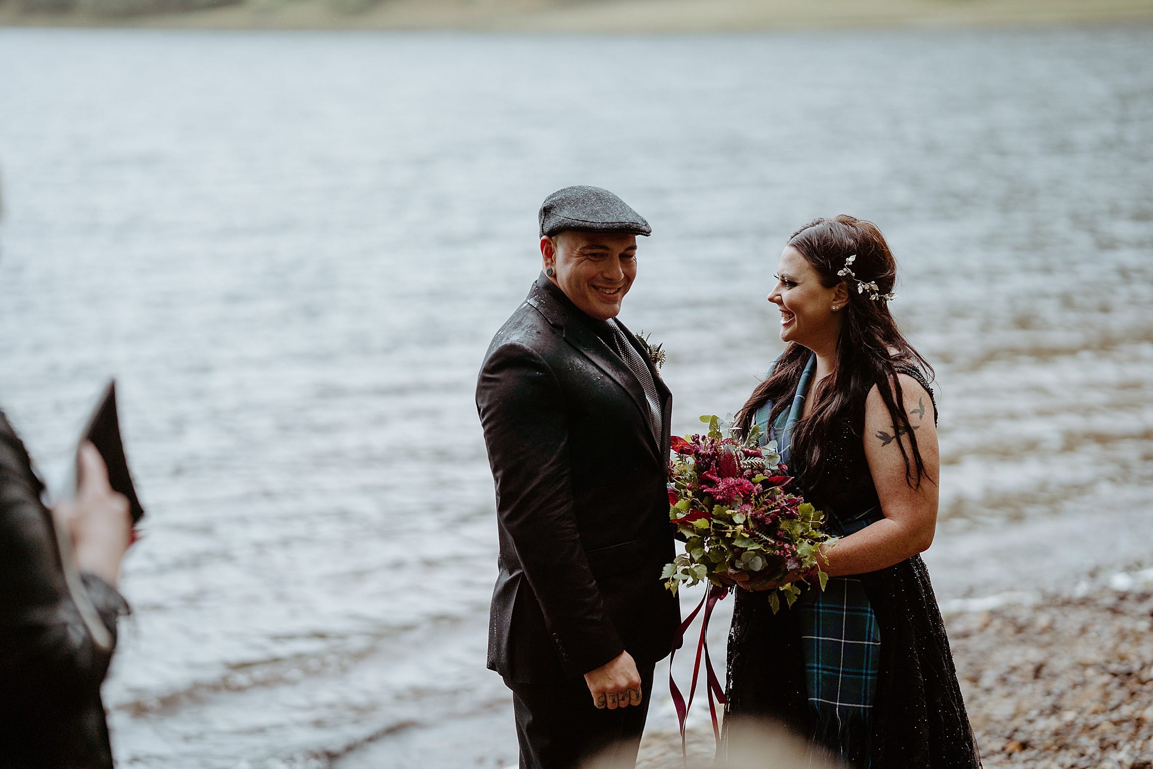 bride and groom smiling as they stand on shore of loch both wearing black and she has bridal bouquet monachyle mhor wedding