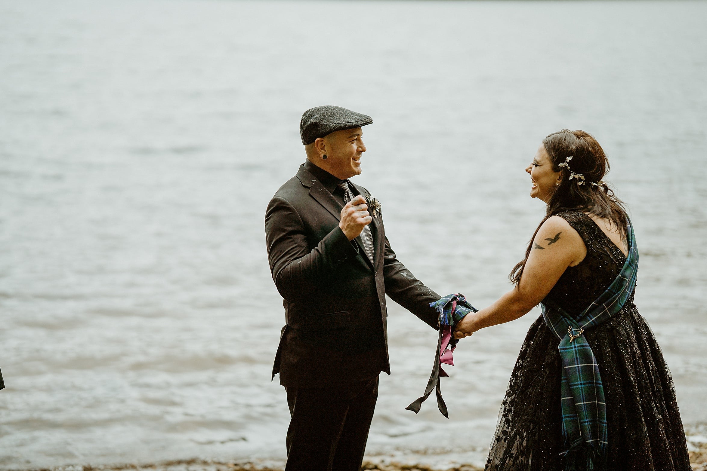 bride and groom both wearing black at shore of loch as they tie the knot with tartan scarf monachyle mhor wedding