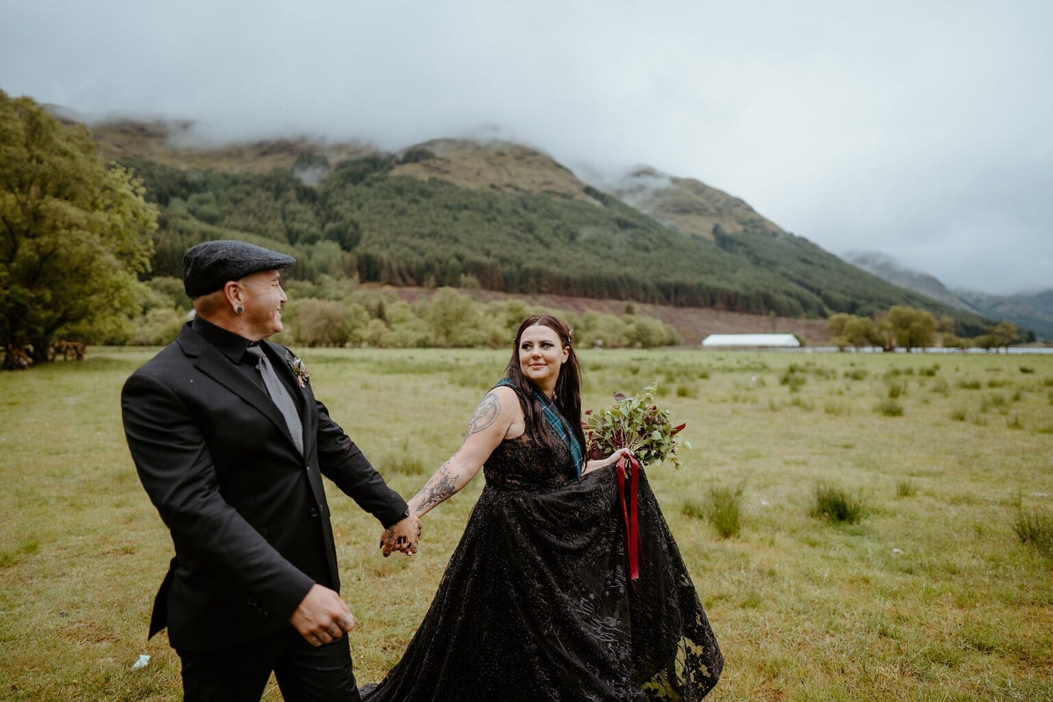 bride and groom both wearing black holding hands as they walk through grass field with green mountains in background monachyle mhor wedding