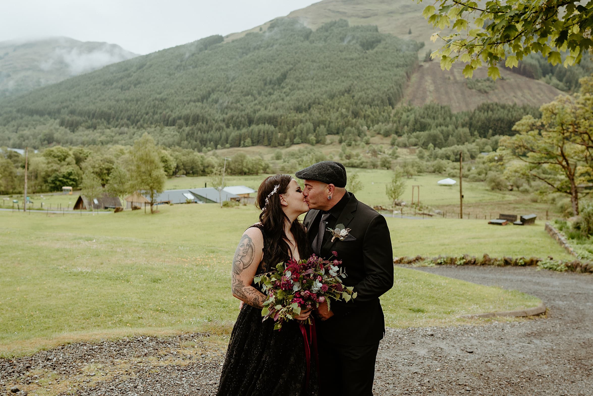 bride and groom both wearing black kiss on country path with grass fields and green mountains in background monachyle mhor wedding