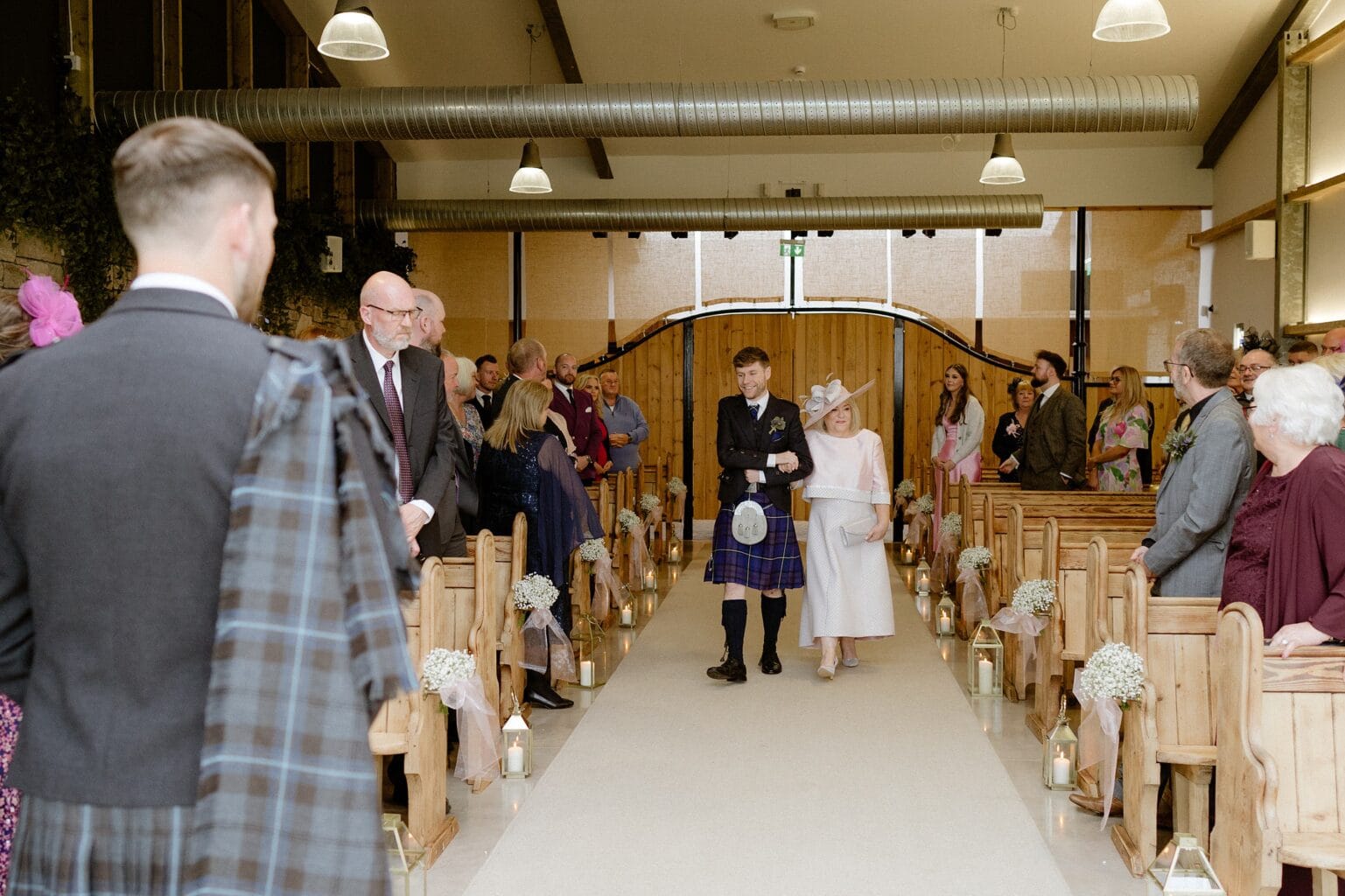 guests watch as the mother of the bride walks down the aisle arm in arm with a groomsman at barn wedding venues west lothian scotland