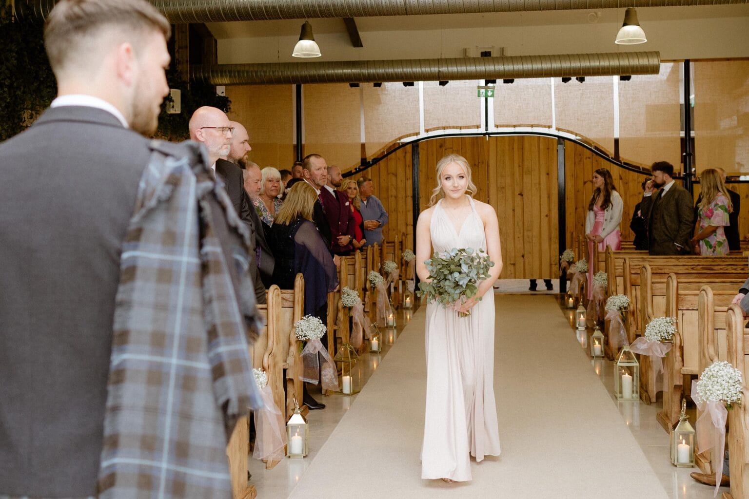 guests stand and watch as a bridesmaid wearing a long pink dress and carrying a greenery bouquet walks down the aisle at barn wedding venues west lothian scotland
