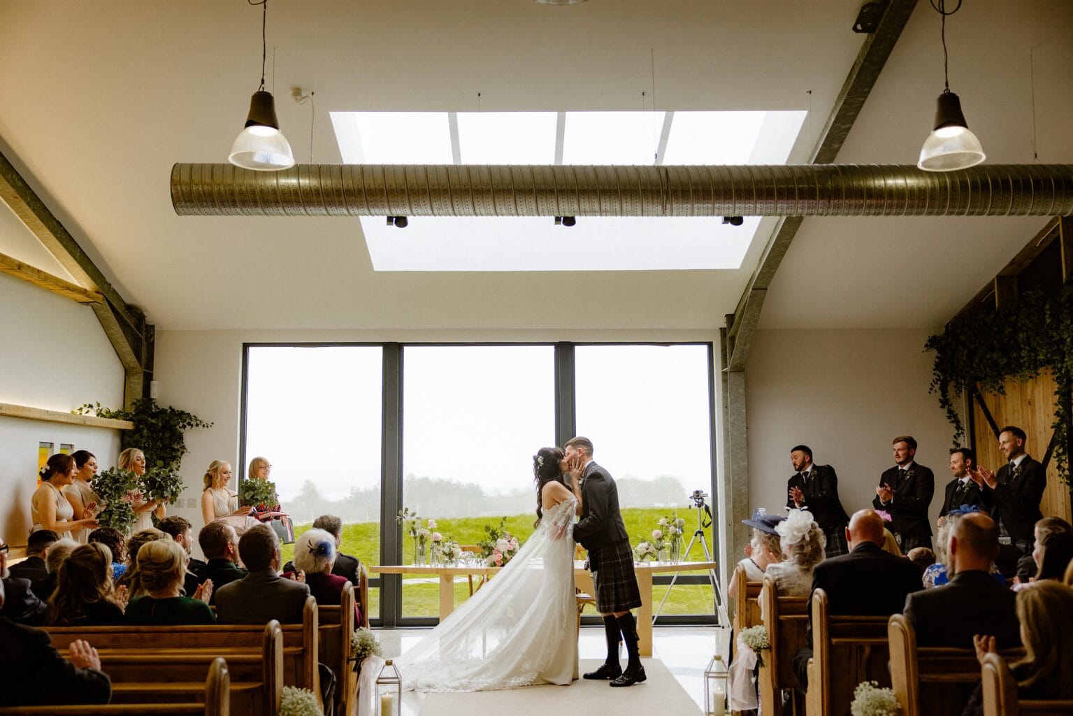guests applaud the bride and groom's first kiss during their wedding ceremony at barn wedding venues west lothian in scotland they are standing in front of a large window with grass and trees visible in the background