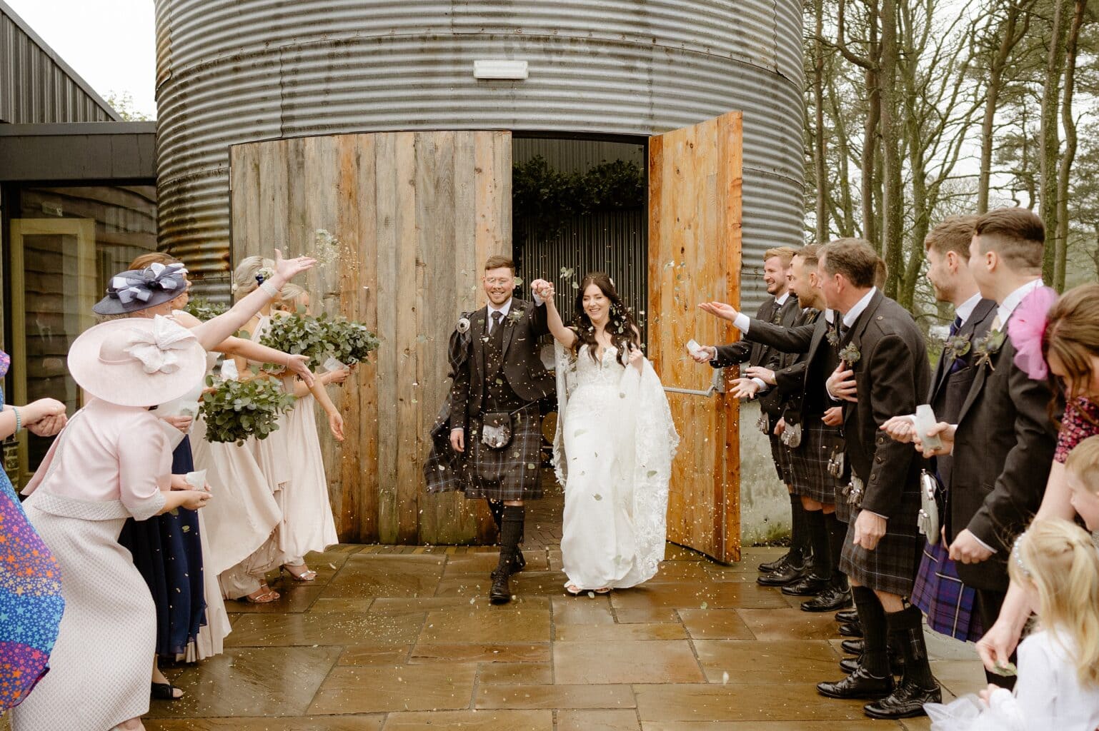 guests toss confetti as the newly married bride and groom exit the barn wedding venues west lothian smiling and holding hands