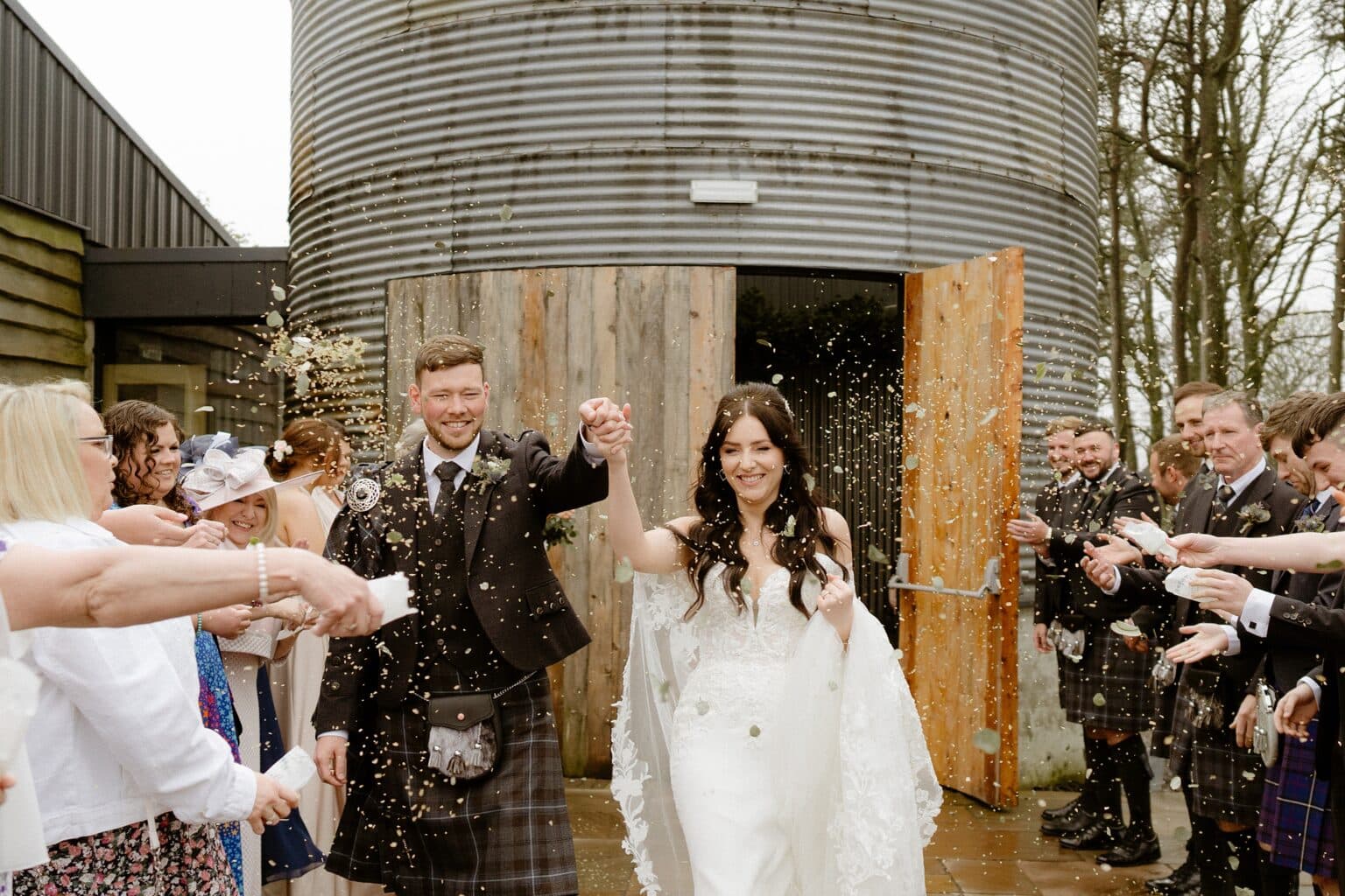 guests toss confetti as the newly married bride and groom exit the barn wedding venues west lothian smiling and holding hands