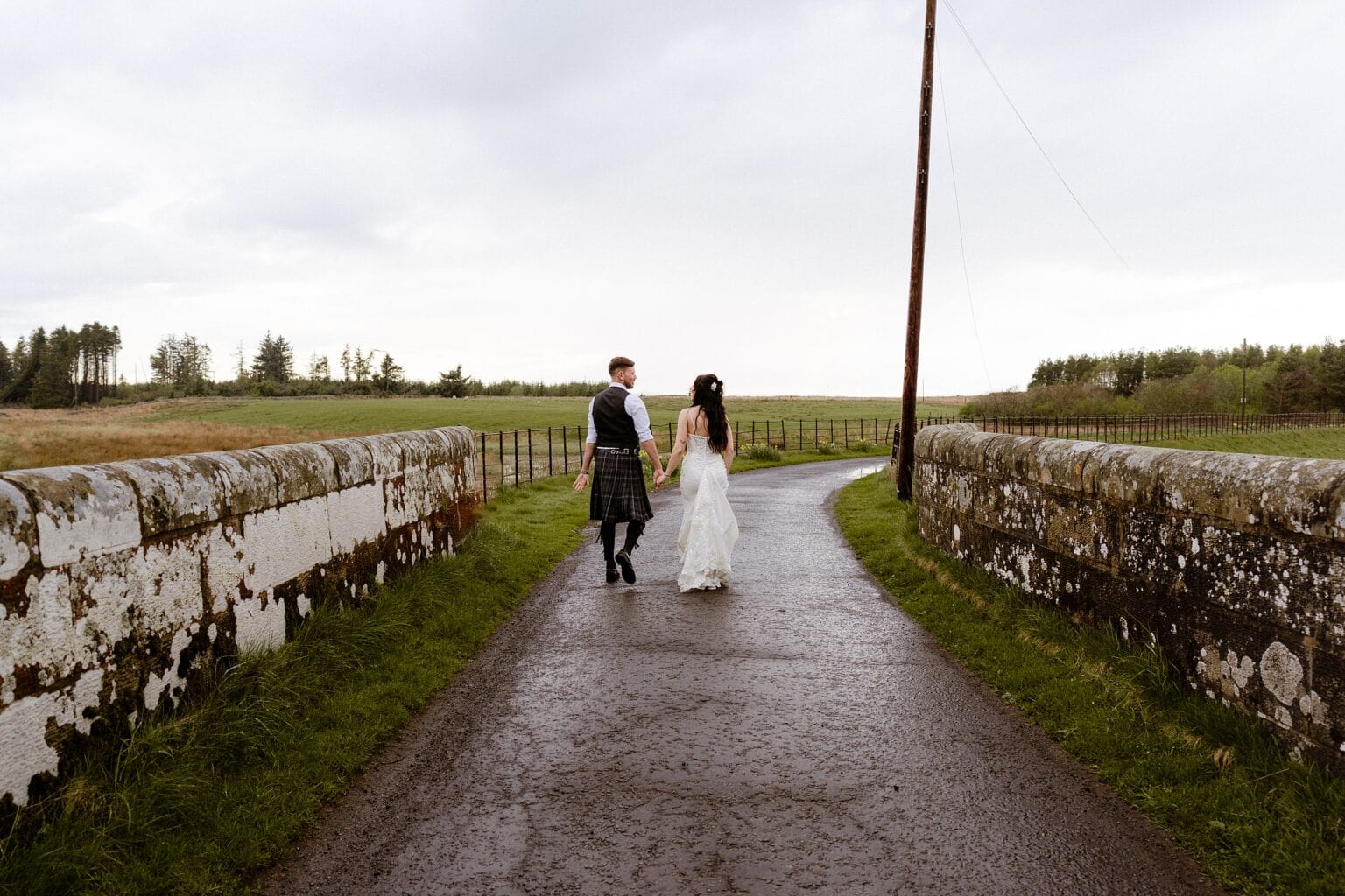 the bride and groom walk along a country path between stone walls with grass trees and sheep in the distance outside their barn wedding venues west lothian scotland