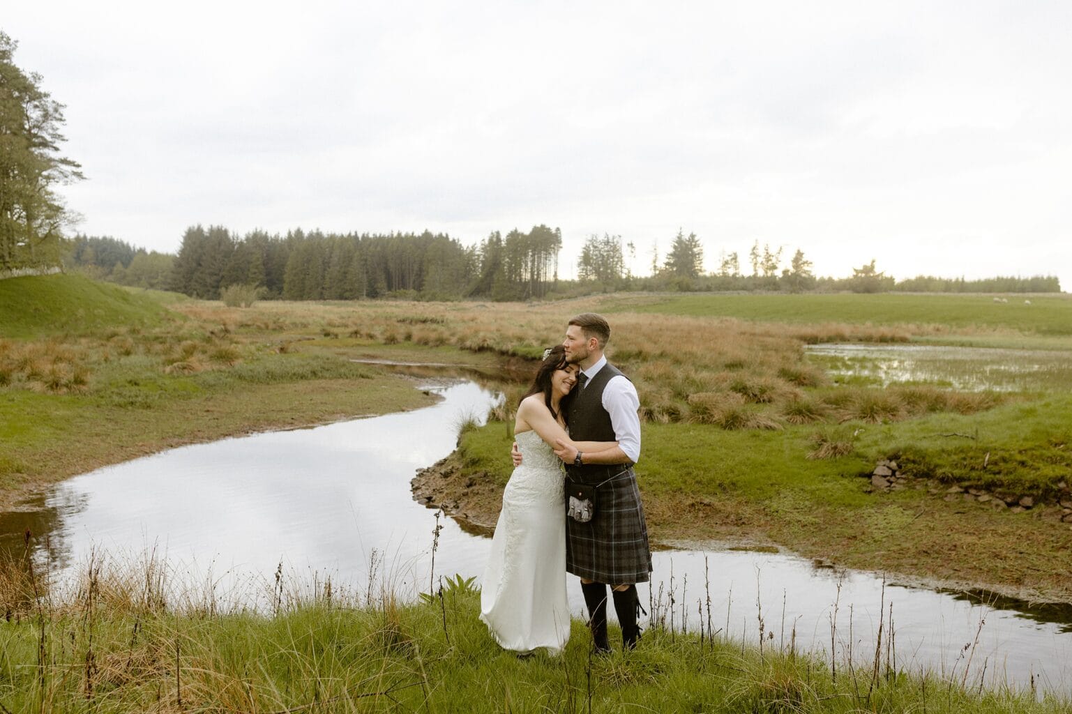 the bride and groom stand in front of a river with their arms around each other with grass and trees visible in the background