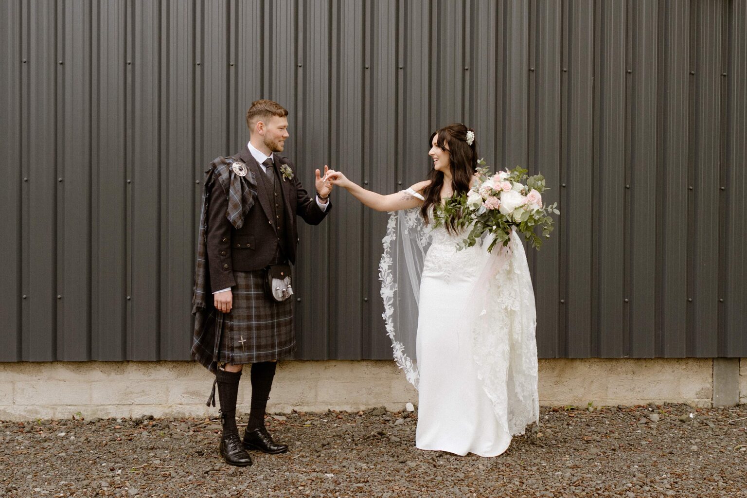 the groom holds the brides hand as they stand in front of a black corrugated iron wall outside their barn wedding venues west lothian in scotland