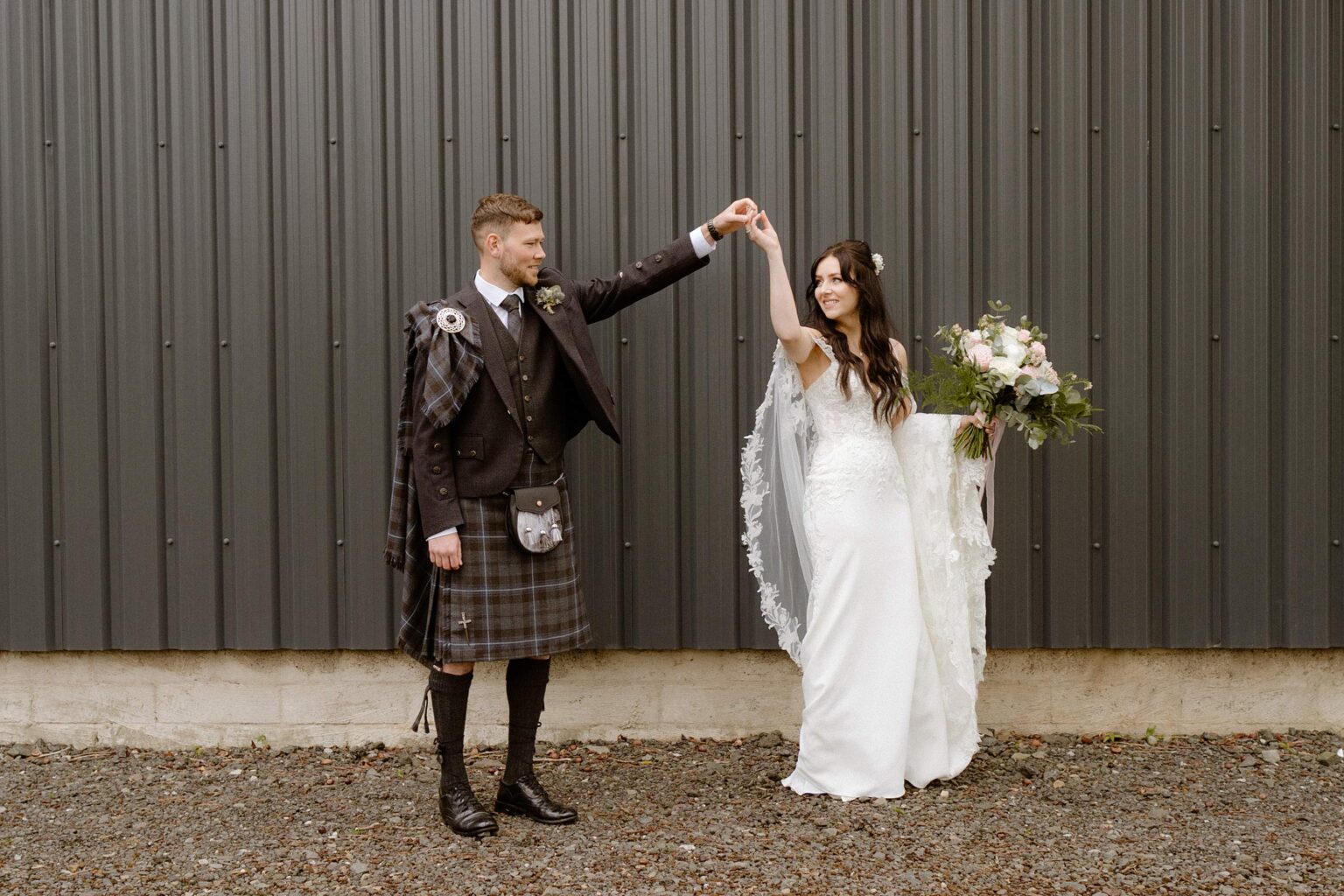 the groom holds the brides hand aloft as she twirls around they are standing in front of a black corrugated iron wall outside their barn wedding venues west lothian in scotland