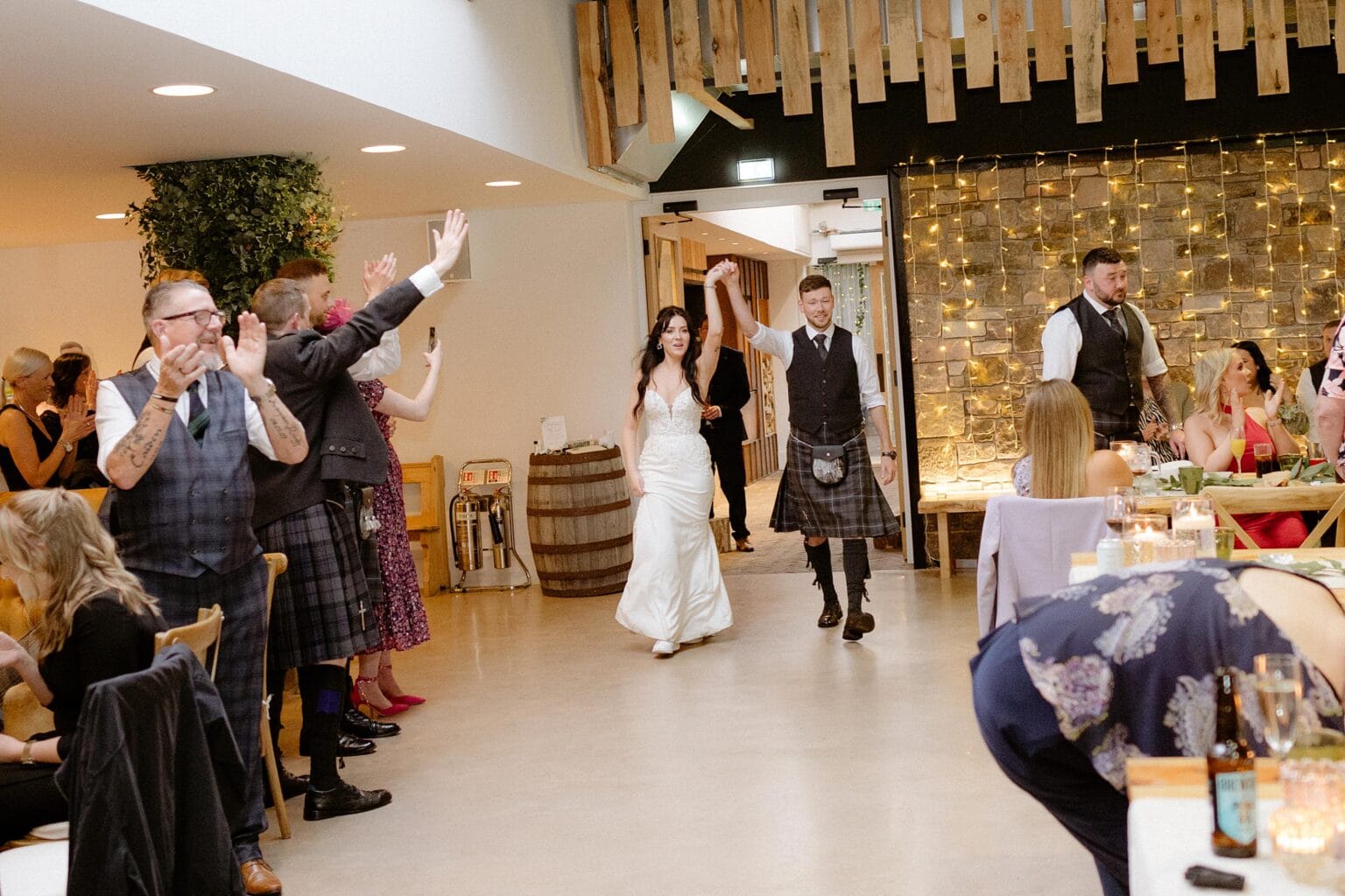 guests applaud as the bride and groom arrive for their wedding dinner at their barn wedding venues west lothian wooden paneling and strings of fairy lights are visible in the background
