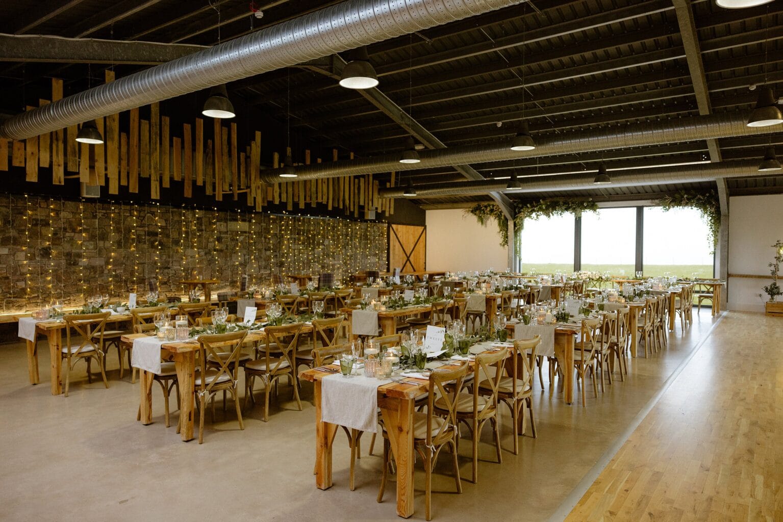 rows of tables set for dinner with glassware candles and greenery at barn wedding venues west lothian a large window wooden paneling and stings of fairy lights are visible in the background