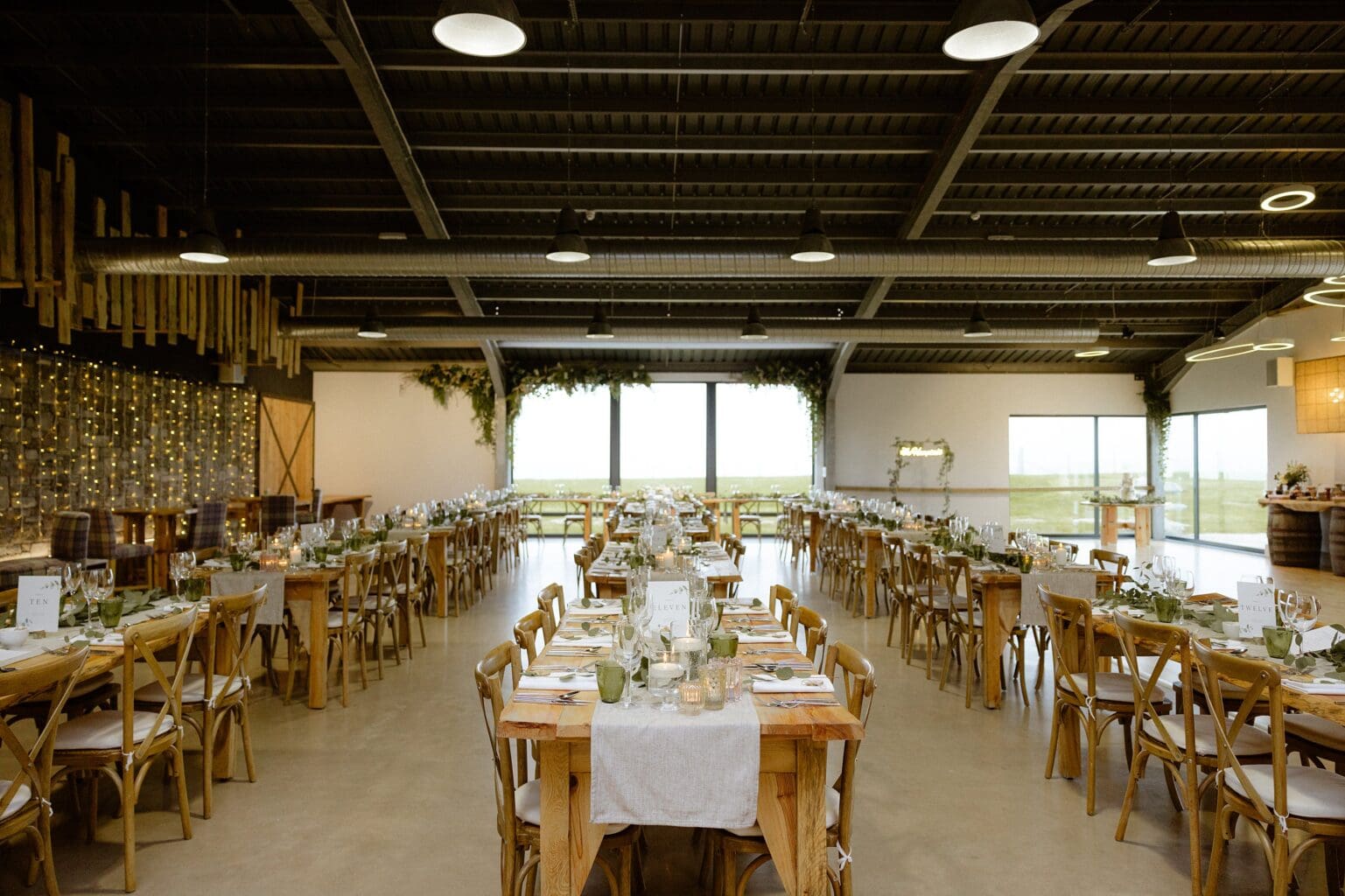 rows of tables set for dinner with glassware candles and greenery at barn wedding venues west lothian a large window wooden paneling and stings of fairy lights are visible in the background