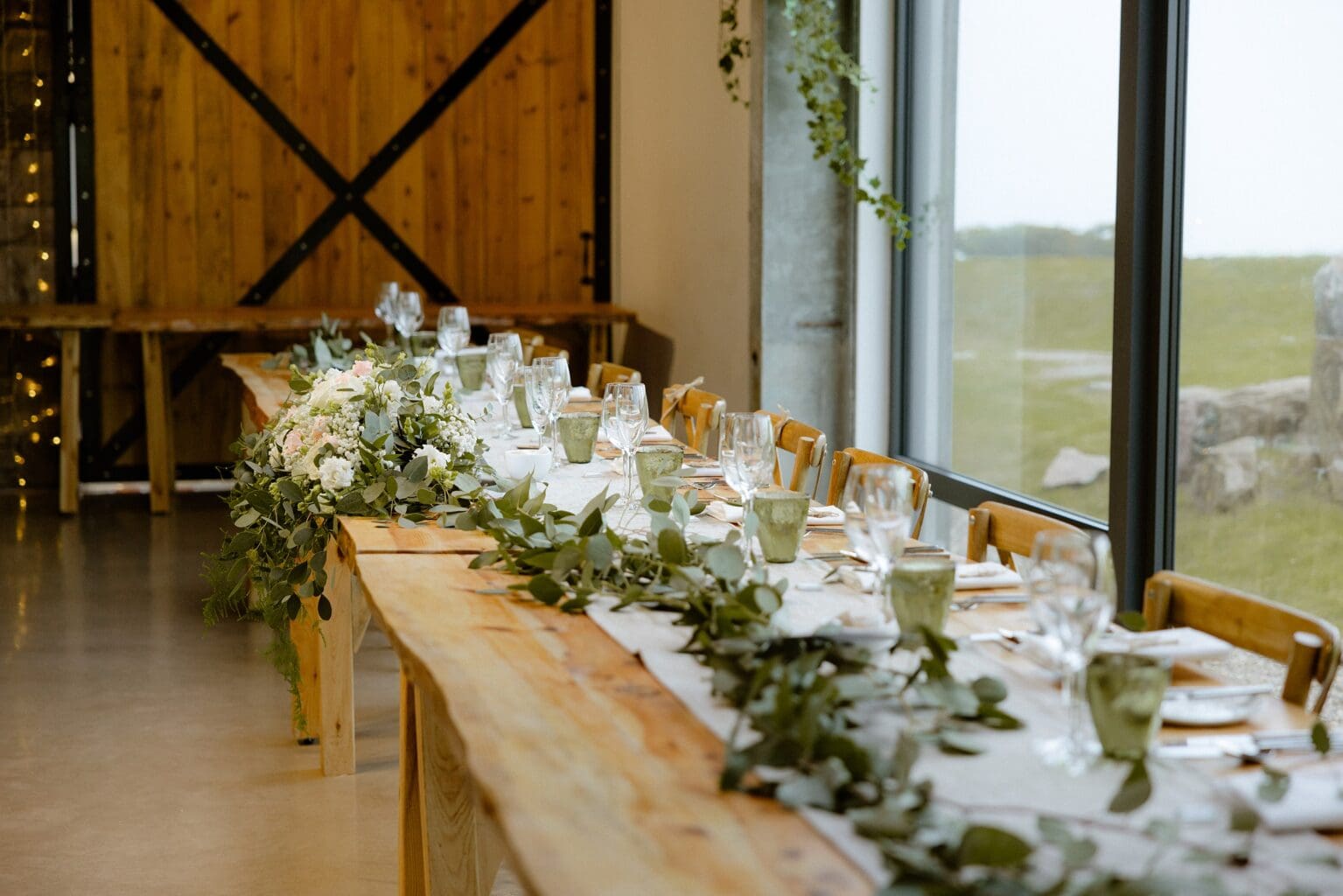 the top table set for dinner with glassware and greenery at barn wedding venues west lothian a large window wooden paneling and stings of fairy lights are visible in the background