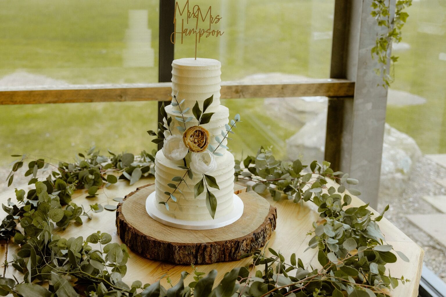a white three tier wedding cake decorated with flowers sits on a wooden base within a greenery wreath in front of a large window
