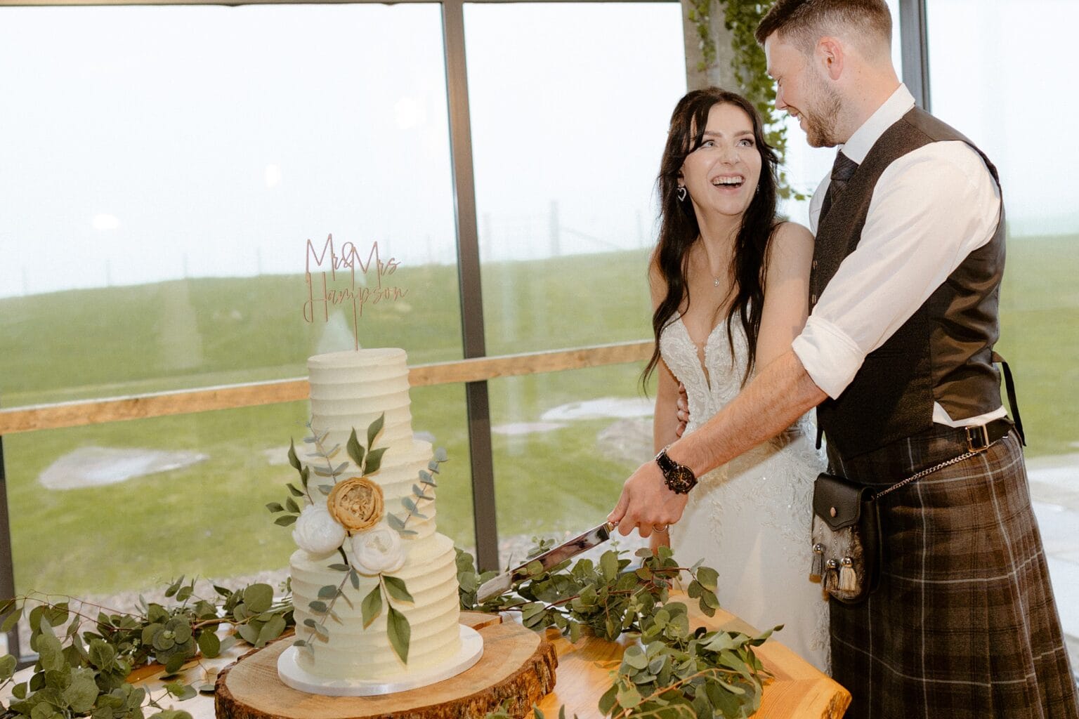 the bride and groom smile as they cut their wedding cake in front of a large window following their wedding ceremony at cairns farm estate kirknewton west lothian