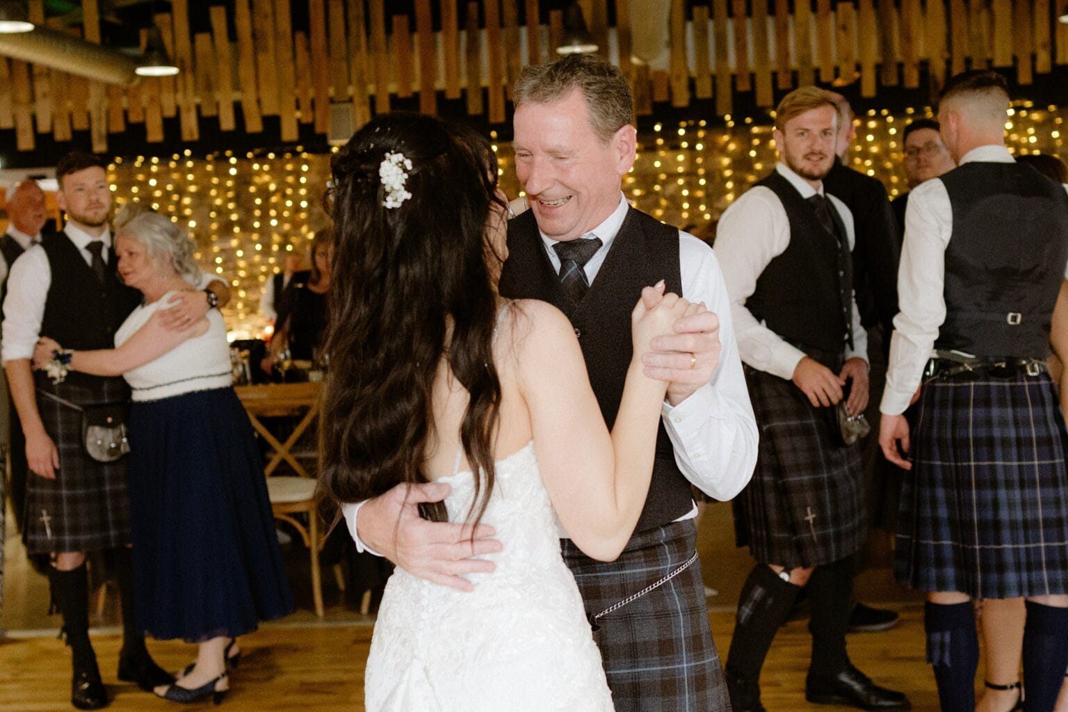 the bride and her father dance together at the evening reception following the wedding ceremony at barn wedding venues west lothian wooden paneling and strings of fairly lights are visible in the background