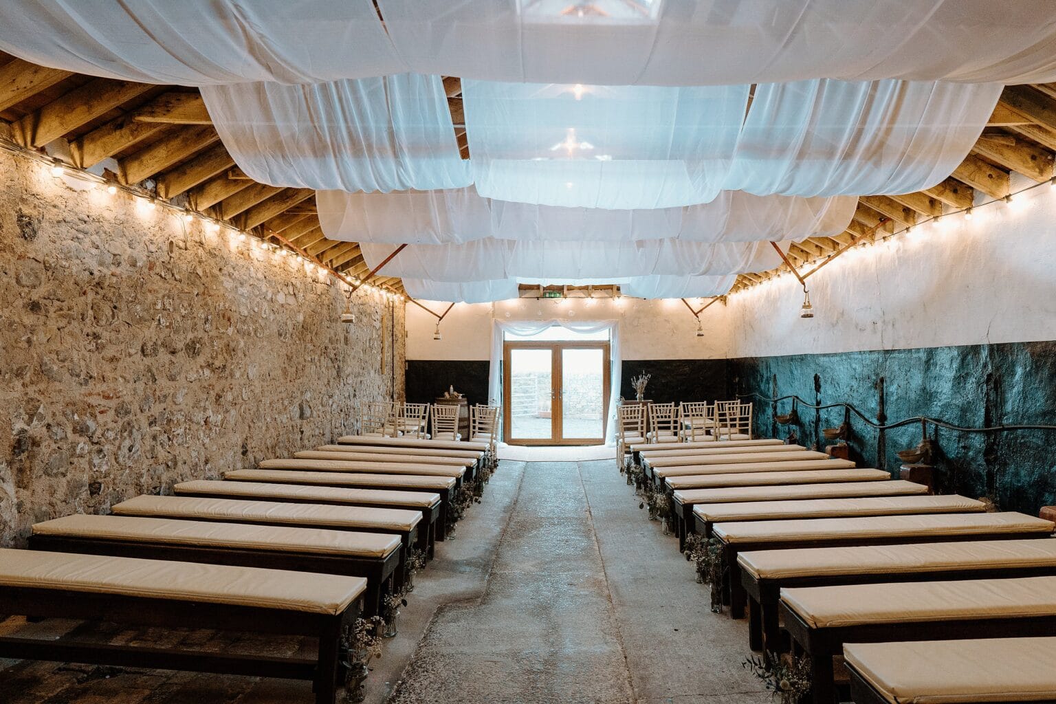 interior inside view of a barn wedding venue in scotland set up for a wedding ceremony with bench seating and white drapes hanging from the ceiling