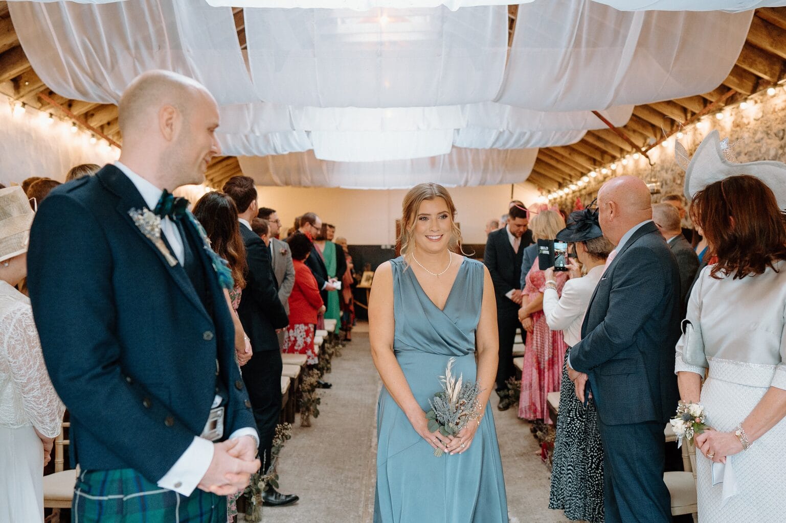 a bridesmaid in a blue dress walks up the aisle as guests look on underneath white drapes hanging from the ceiling in one of the best farm wedding venues in scotland