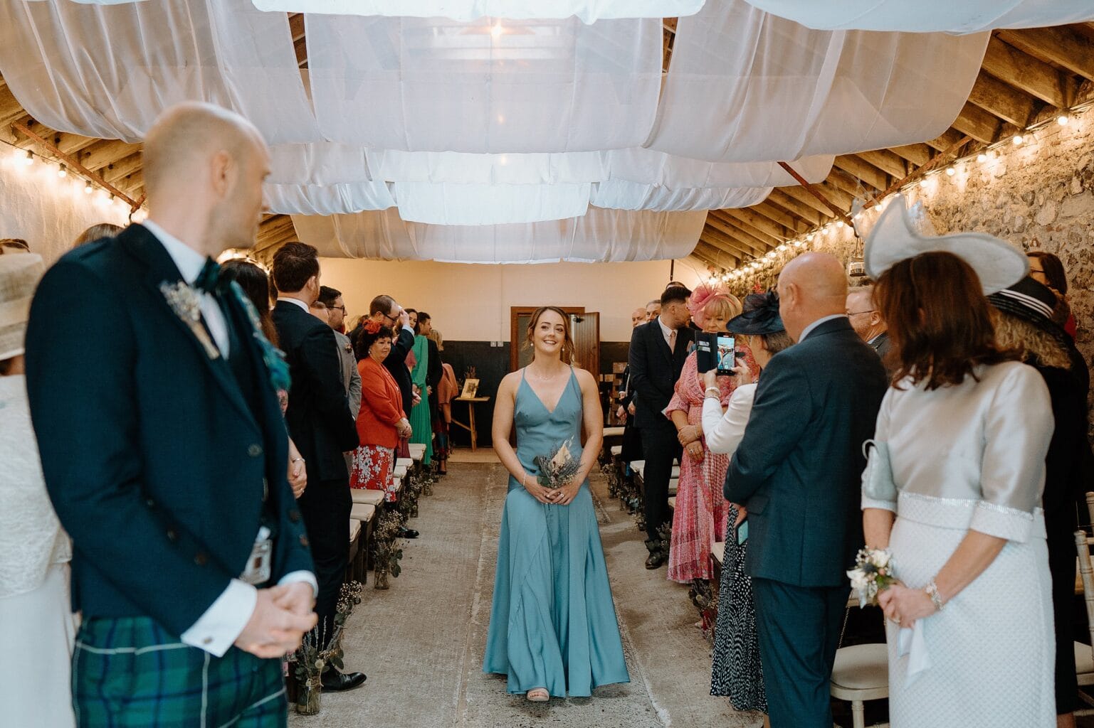 a bridesmaid in a blue dress walks up the aisle as guests look on underneath white drapes hanging from the ceiling in one of the best farm wedding venues in scotland