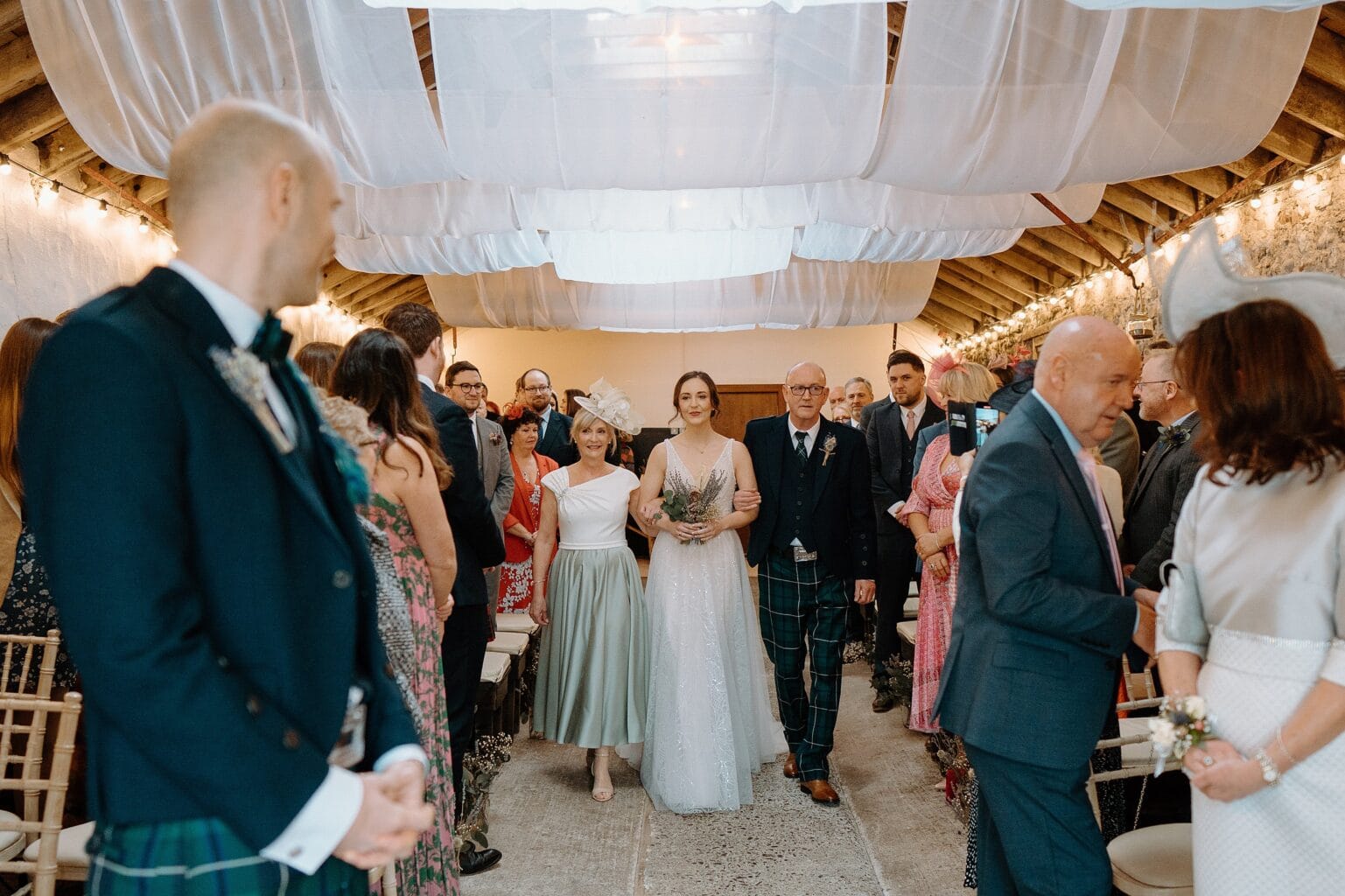 the bride walks down the aisle arm in arm with her parents as guests and the groom look on at one of the most unique farm wedding venues in scotland