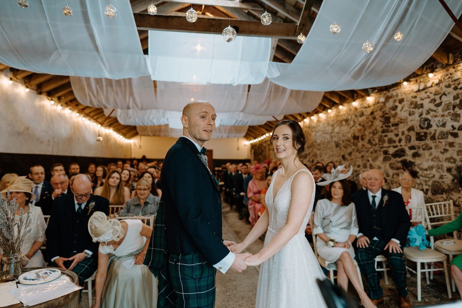 seating guests look on as the bride and groom hold hands beneath decorative white drapes during their wedding ceremony at one of the most unusual farm wedding venues in scotland