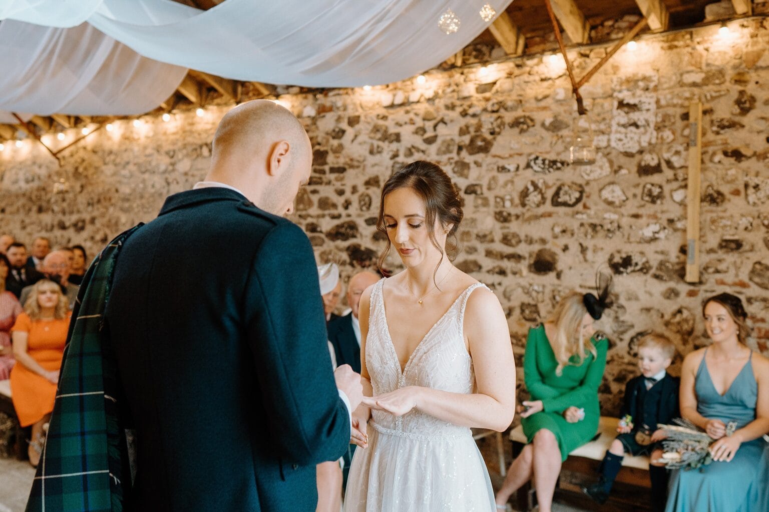the groom places the wedding ring on the bride's finger during their wedding ceremony in a barn wedding venue near glasgow scotland
