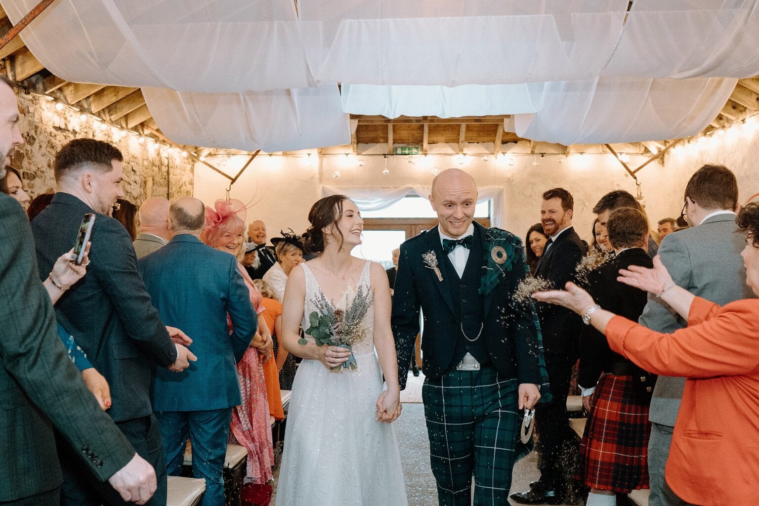 a guest tosses dried flower confetti as the bride and groom walk down the aisle holding hands following their wedding ceremony at a barn wedding venue near glasgow scotland