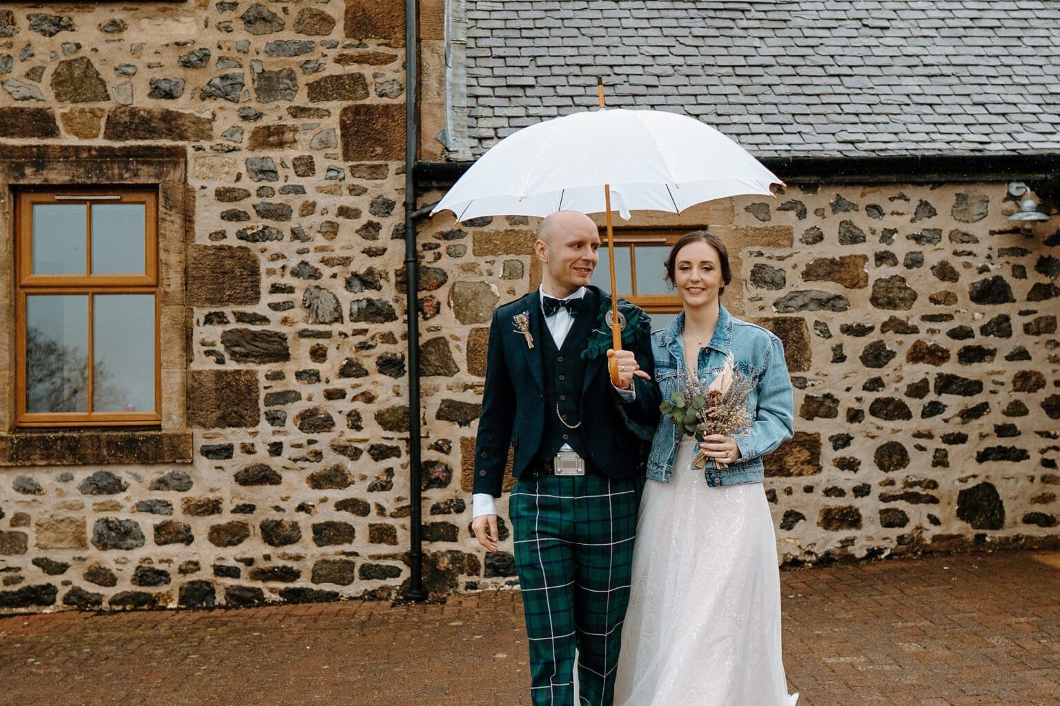 the bride is wearing a blue denim jacket and holding her dried flower bouquet as she and the groom stand underneath a white umbrella in front of a stone walled barn at their farm wedding venues scotland