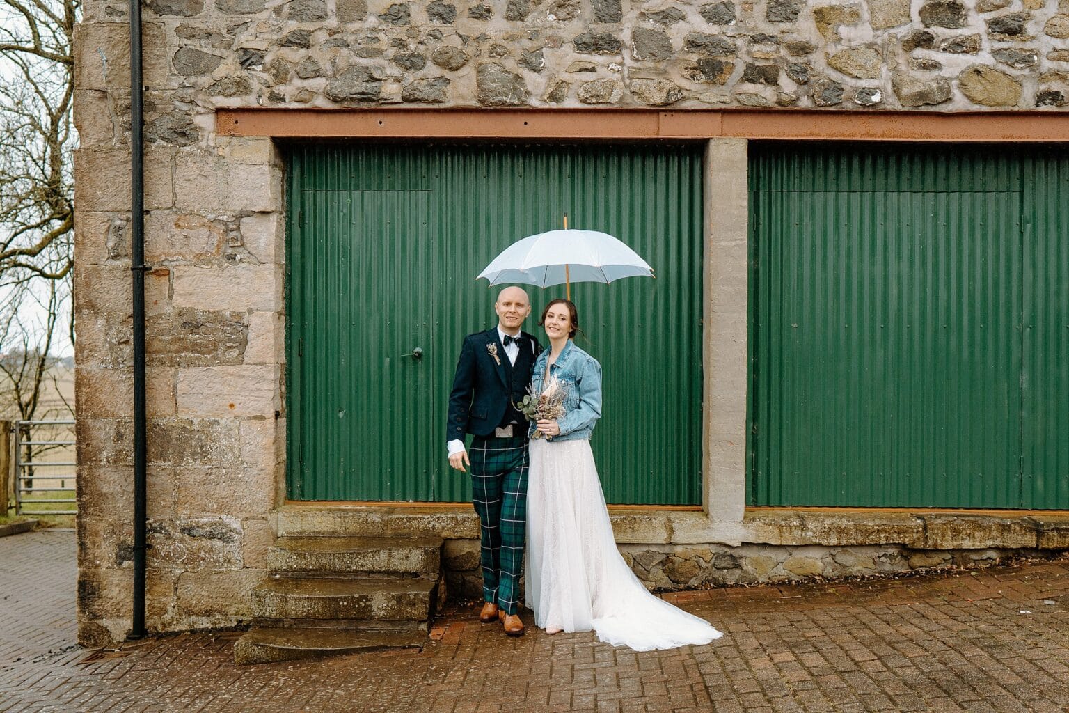 the bride is wearing a blue denim jacket and holding her dried flower bouquet as she and the groom stand underneath a white umbrella in front of green barn doors at their farm wedding venues scotland