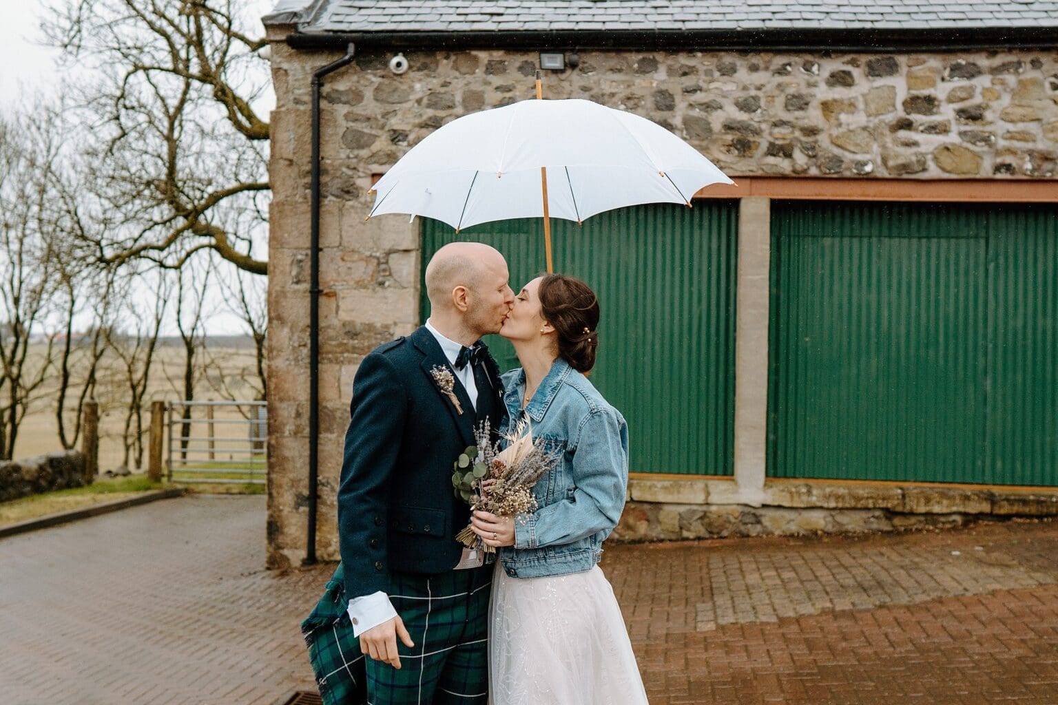 the bride is wearing a blue denim jacket and holding her dried flower bouquet as she kisses the groom underneath a white umbrella outside their farm wedding venues scotland