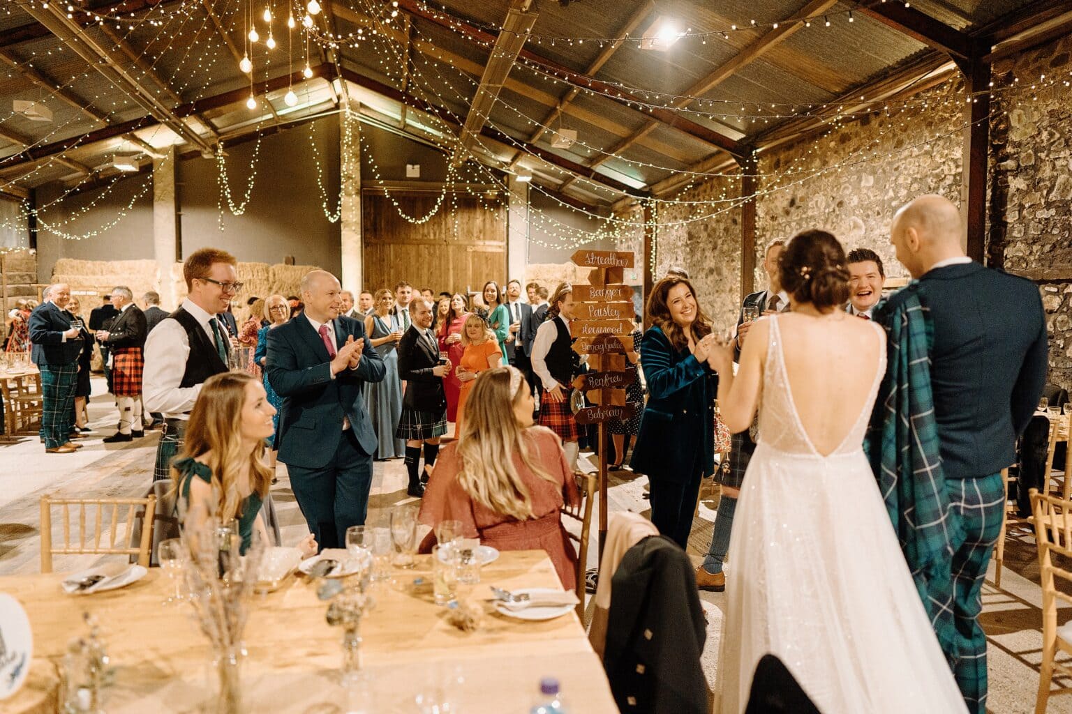 guests stand and applaud beneath festoon lighting in a room set for dinner as the bride and groom enter following their wedding ceremony at their farm wedding venues scotland