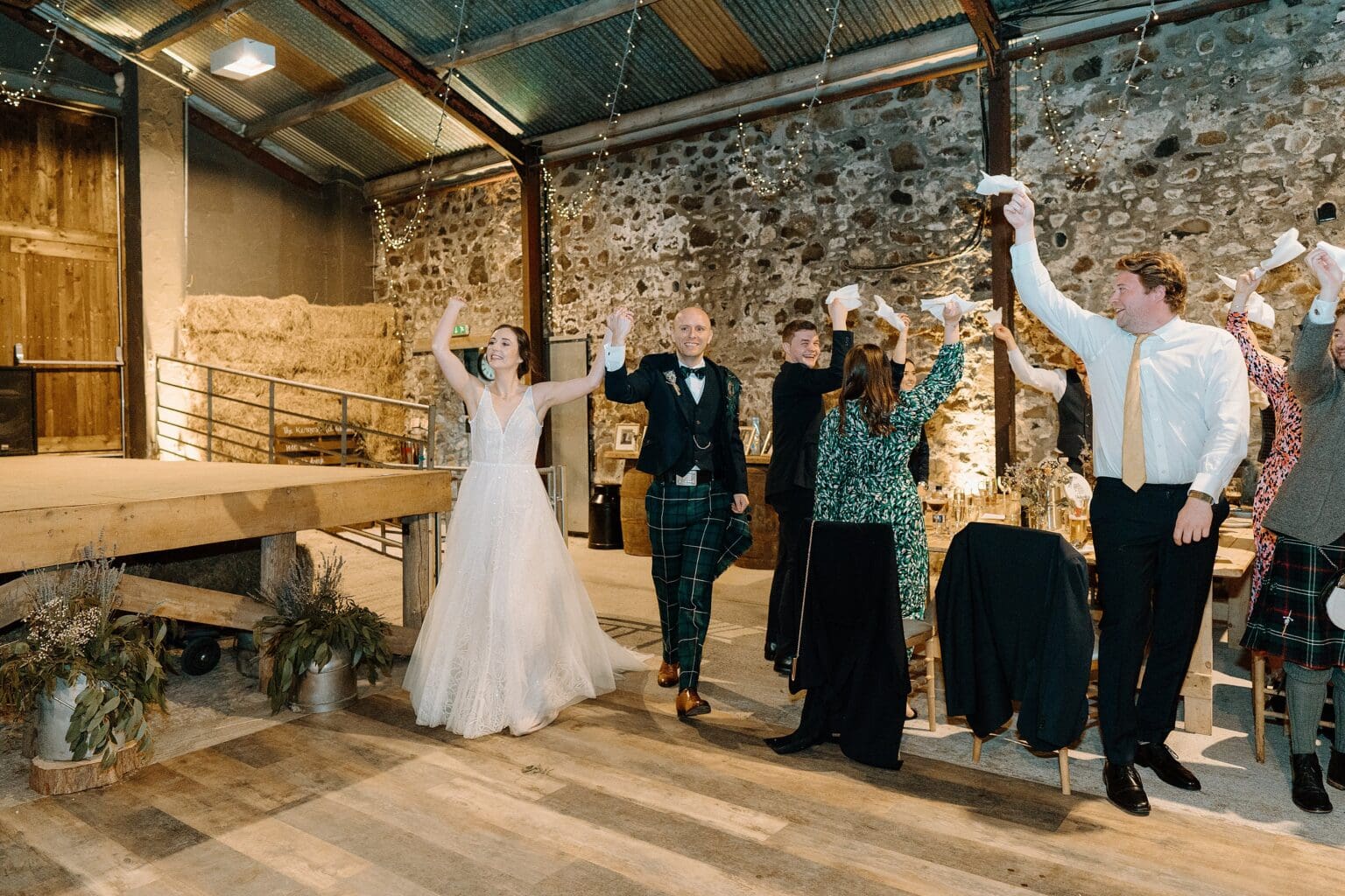in a stone walled room with festoon lighting and hay bales in the background guests stand and wave white napkins as the bride and groom arrive for their wedding dinner at their barn wedding venue near glasgow scotland